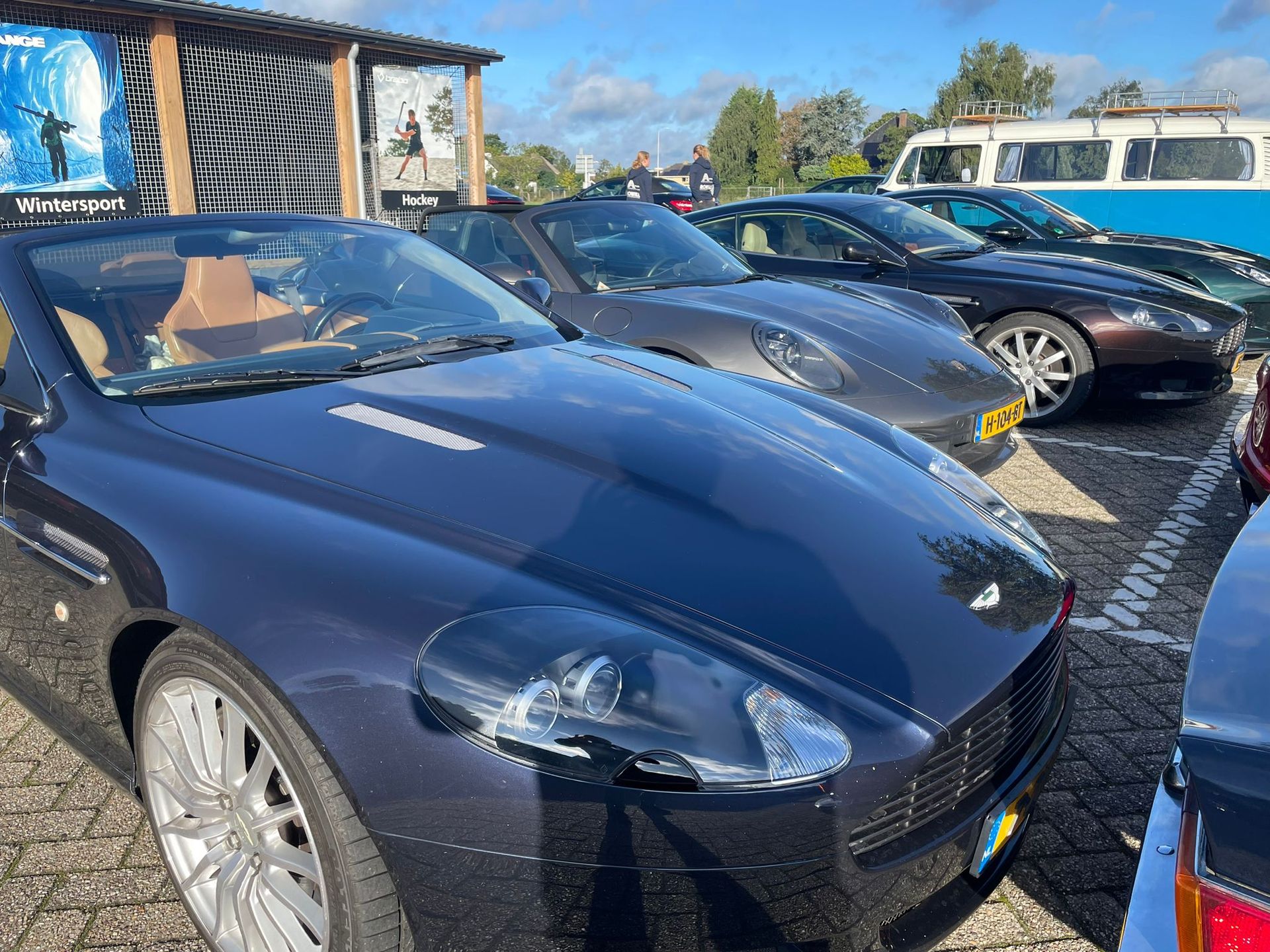 Er stonden diverse luxeauto's buiten geparkeerd, waaronder een blauwe Aston Martin, een zilveren Porsche en een donkerkleurige coupé.