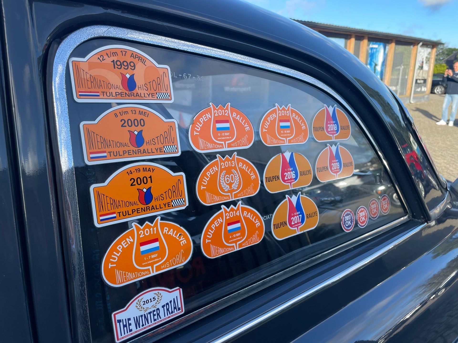 Close-up van een autoruit volgeplakt met oranje en bruine rallystickers; een Nederlandse vlag is te zien.