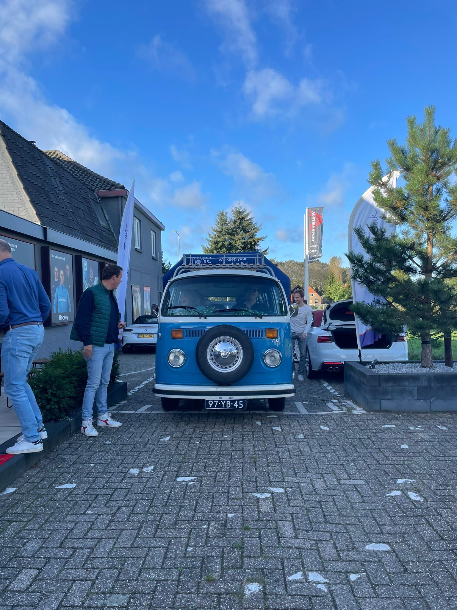 Een blauwe VW-bus staat geparkeerd op een straat met kinderkopjes. Twee mensen staan ​​bij het voertuig, met gebouwen en de lucht op de achtergrond.