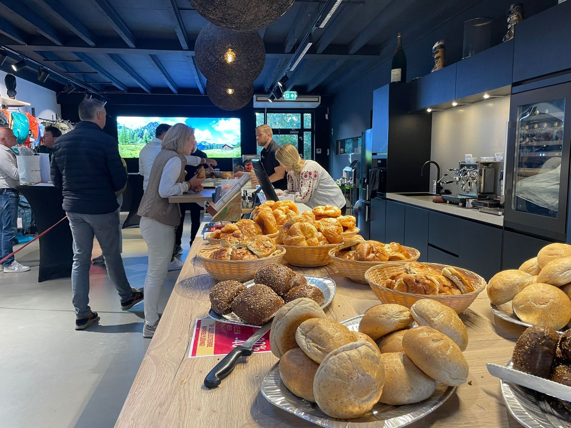 Mensen bij een buffet met brood, gebak en een scherm op de achtergrond. Interieur.