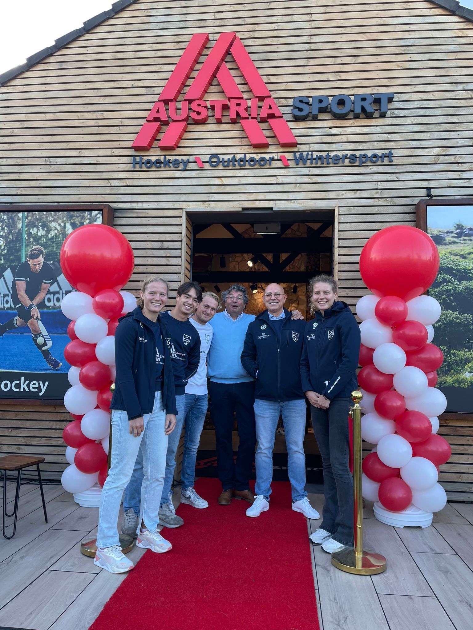 Een groep mensen voor de Austria Sport-winkel, met een rode loper, ballonnen, een houten gevel en een bord met de merknaam.