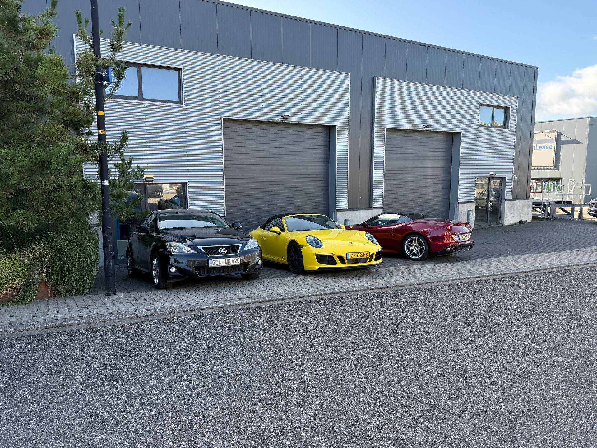 Drie cabriolets staan geparkeerd voor een grijs industrieel gebouw: een zwarte, een gele en een rode.