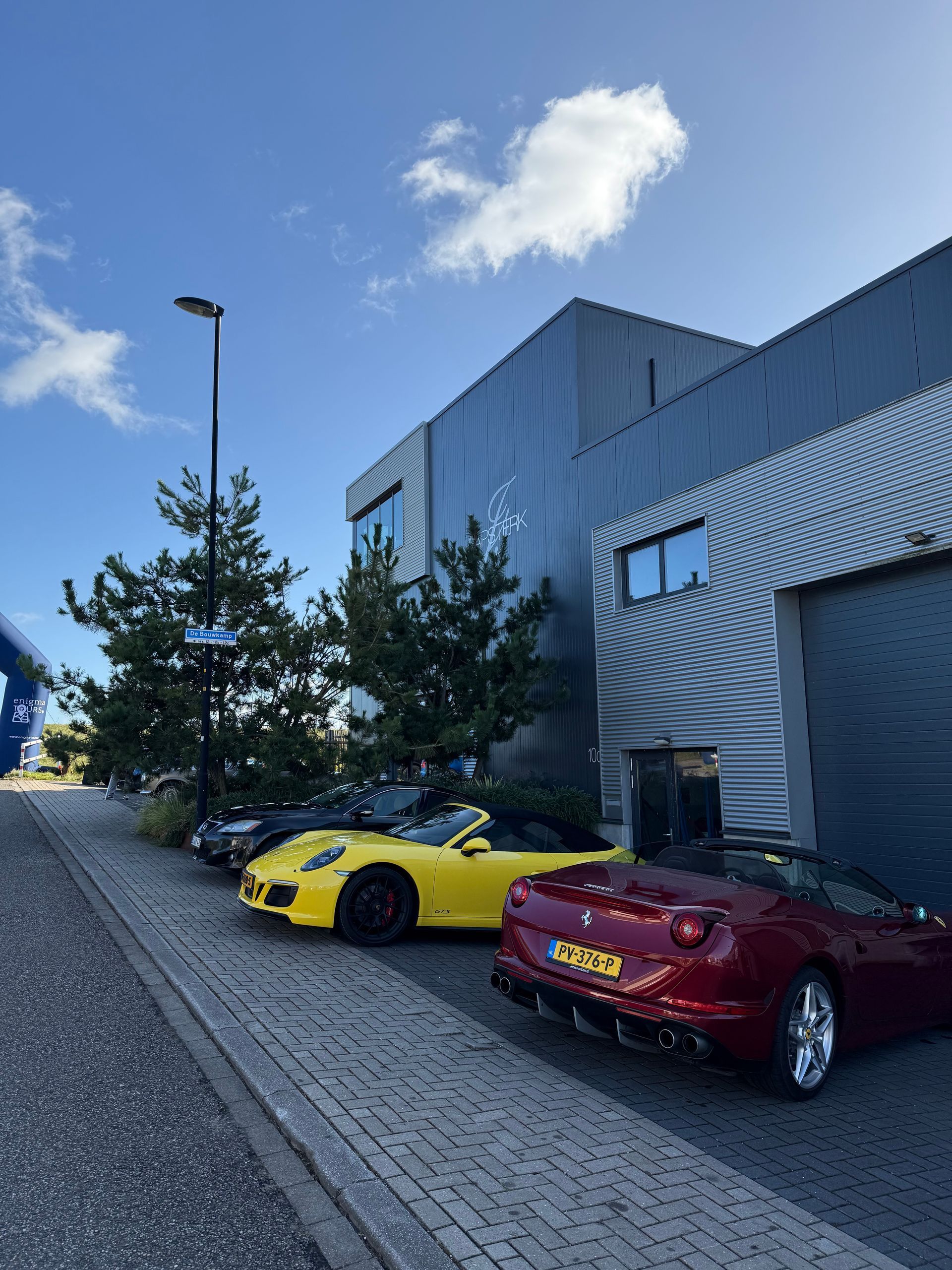 Op een zonnige dag staan er auto's geparkeerd voor een modern gebouw. Gele, rode en zwarte cabriolets zijn zichtbaar.