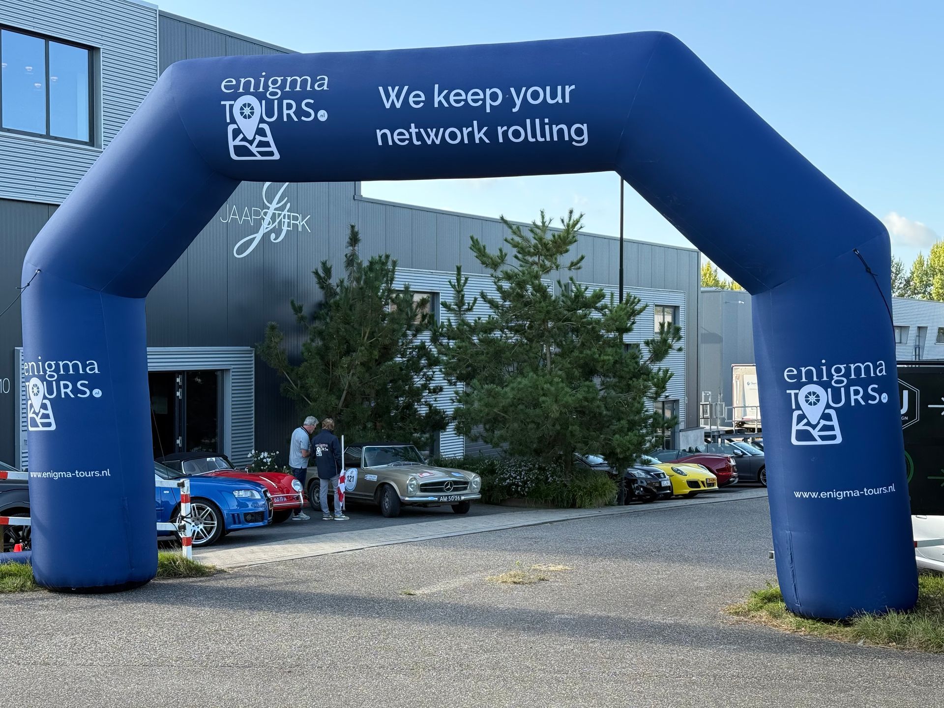 Blauwe opblaasbare boog voor Enigma Tours met auto's geparkeerd voor een gebouw.