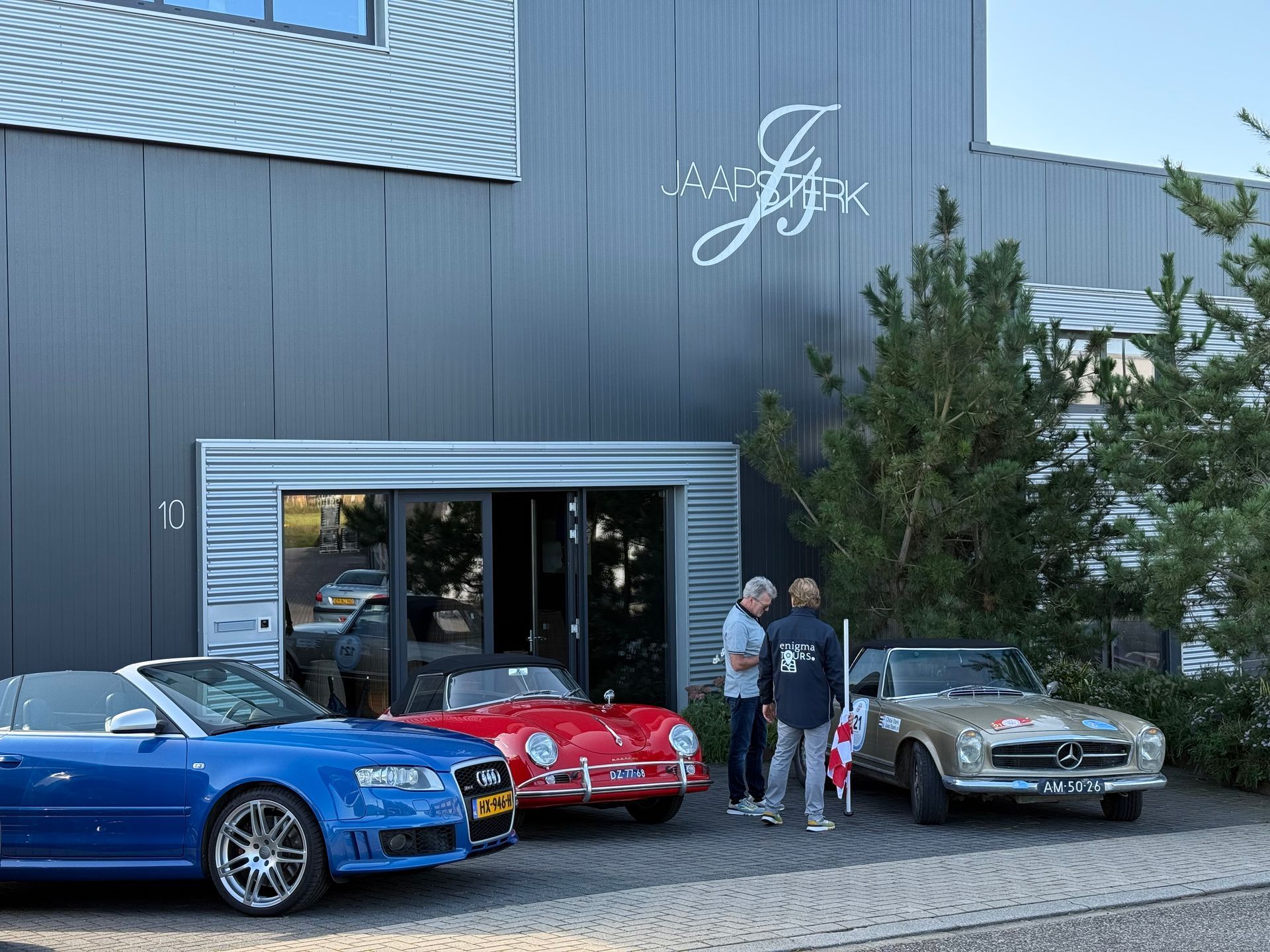 Drie klassieke auto's staan geparkeerd voor een gebouw; mensen staan eromheen.