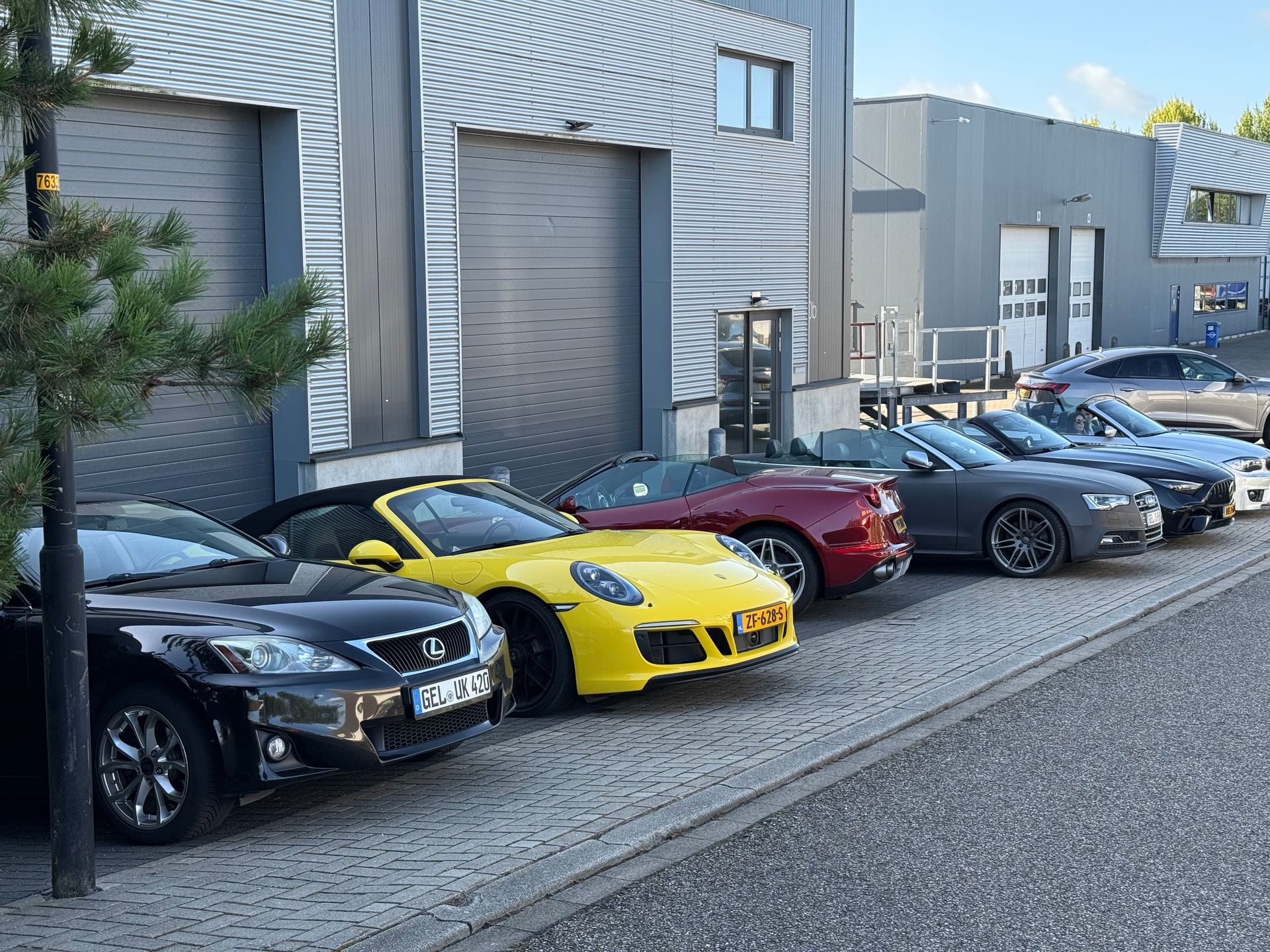 Een rij cabriolets staat geparkeerd voor een grijs gebouw: Lexus, Porsche, Corvette, BMW en andere merken.
