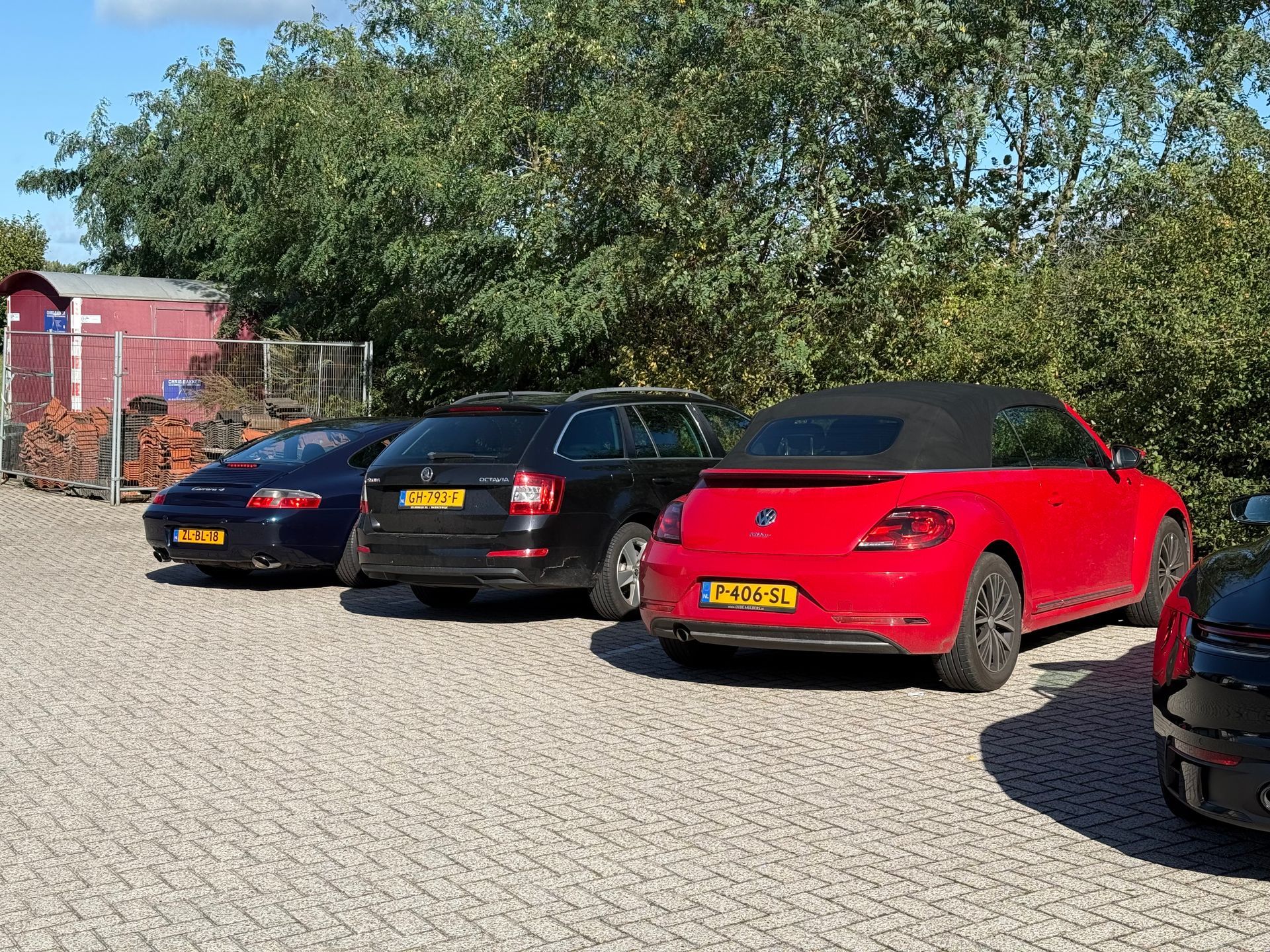 Auto's geparkeerd op een grindterrein: een rode cabriolet, een zwarte stationwagen en een blauwe sedan. Groene bomen en een rode schuur op de achtergrond.
