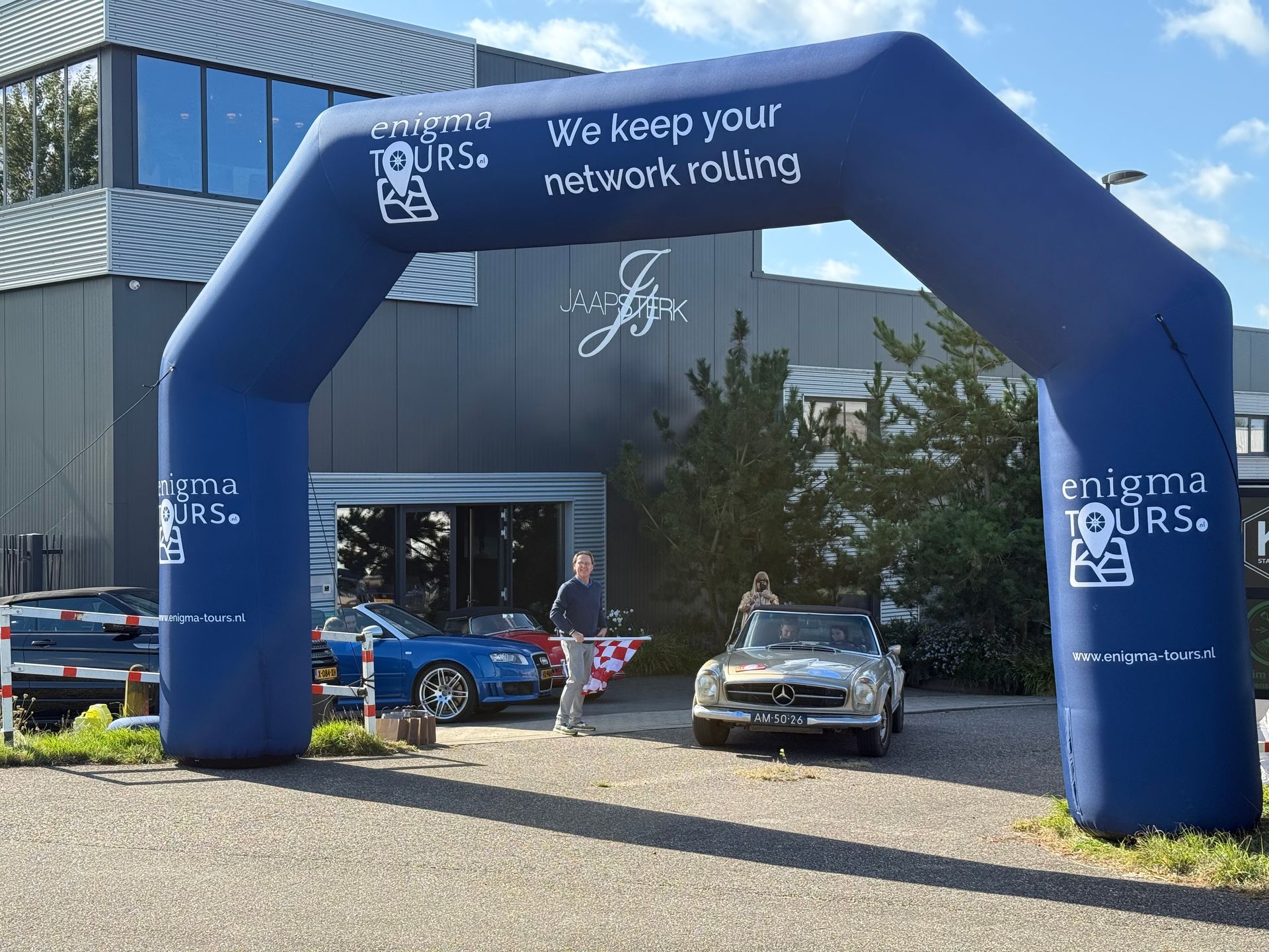Blauwe opblaasbare boog op een evenement. Auto's en mensen zijn zichtbaar. Tekst: