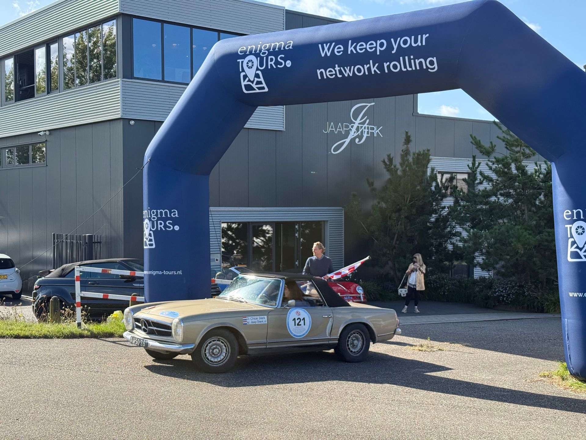 Een beige cabriolet rijdt onder een blauwe boog door, terwijl mensen bij een gebouw met vlaggen zwaaien.