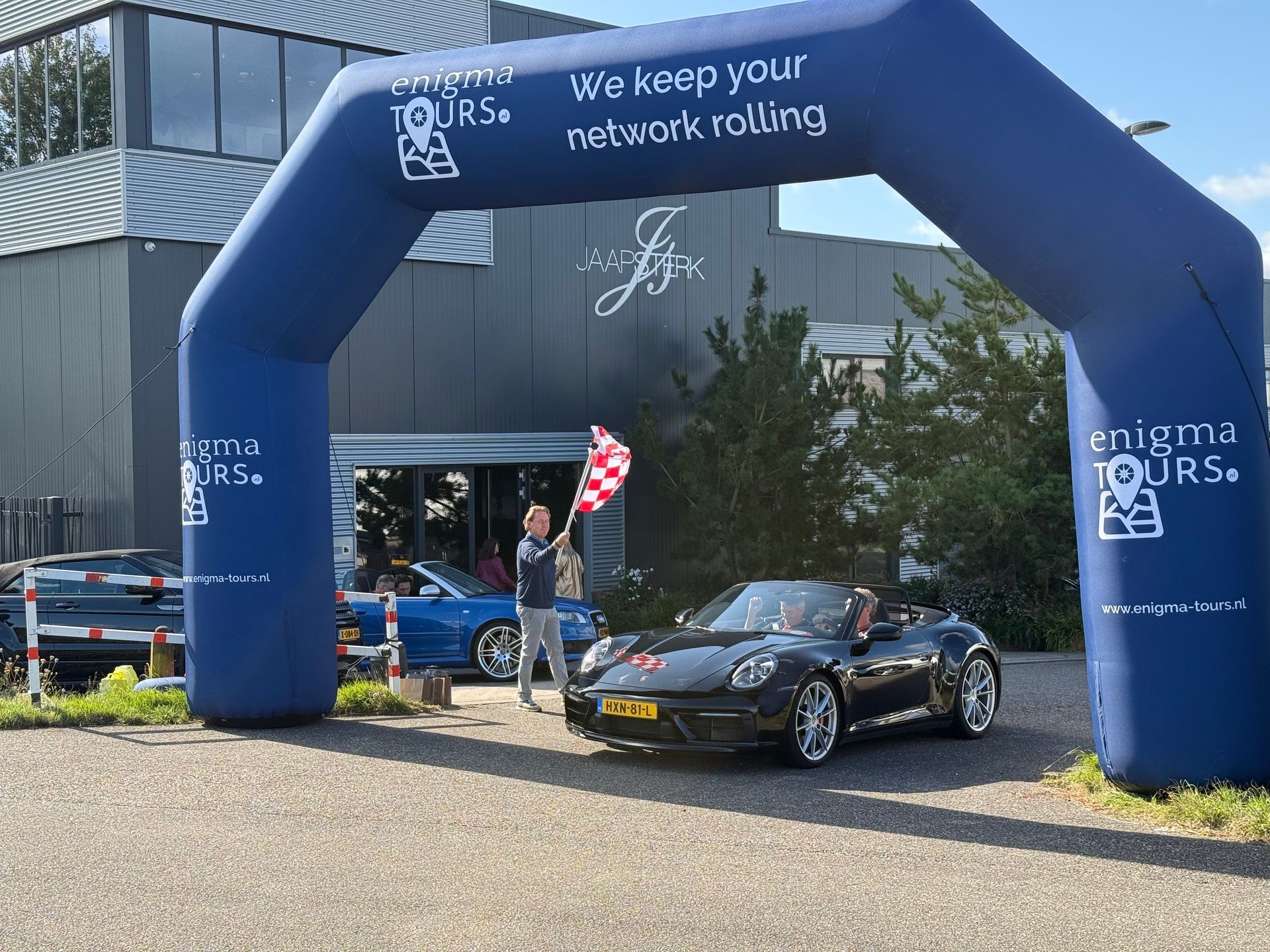 Een zwarte sportwagen rijdt onder een opblaasbare boog door; een man zwaait met een geblokte vlag.
