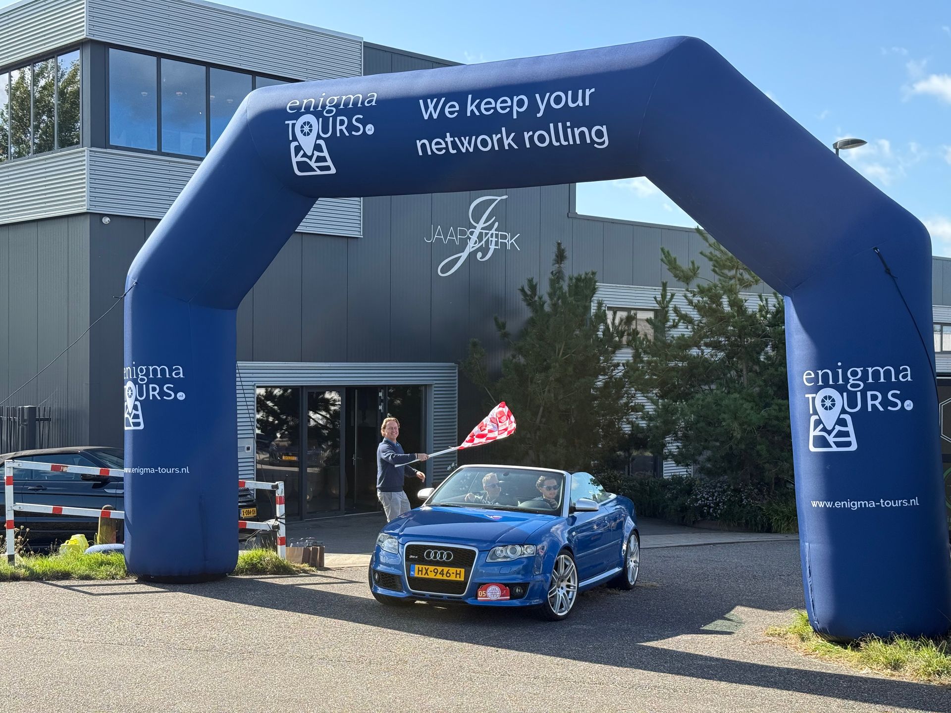 Een blauwe cabriolet rijdt onder een opblaasbare boog door; iemand zwaait met een geblokte vlag.