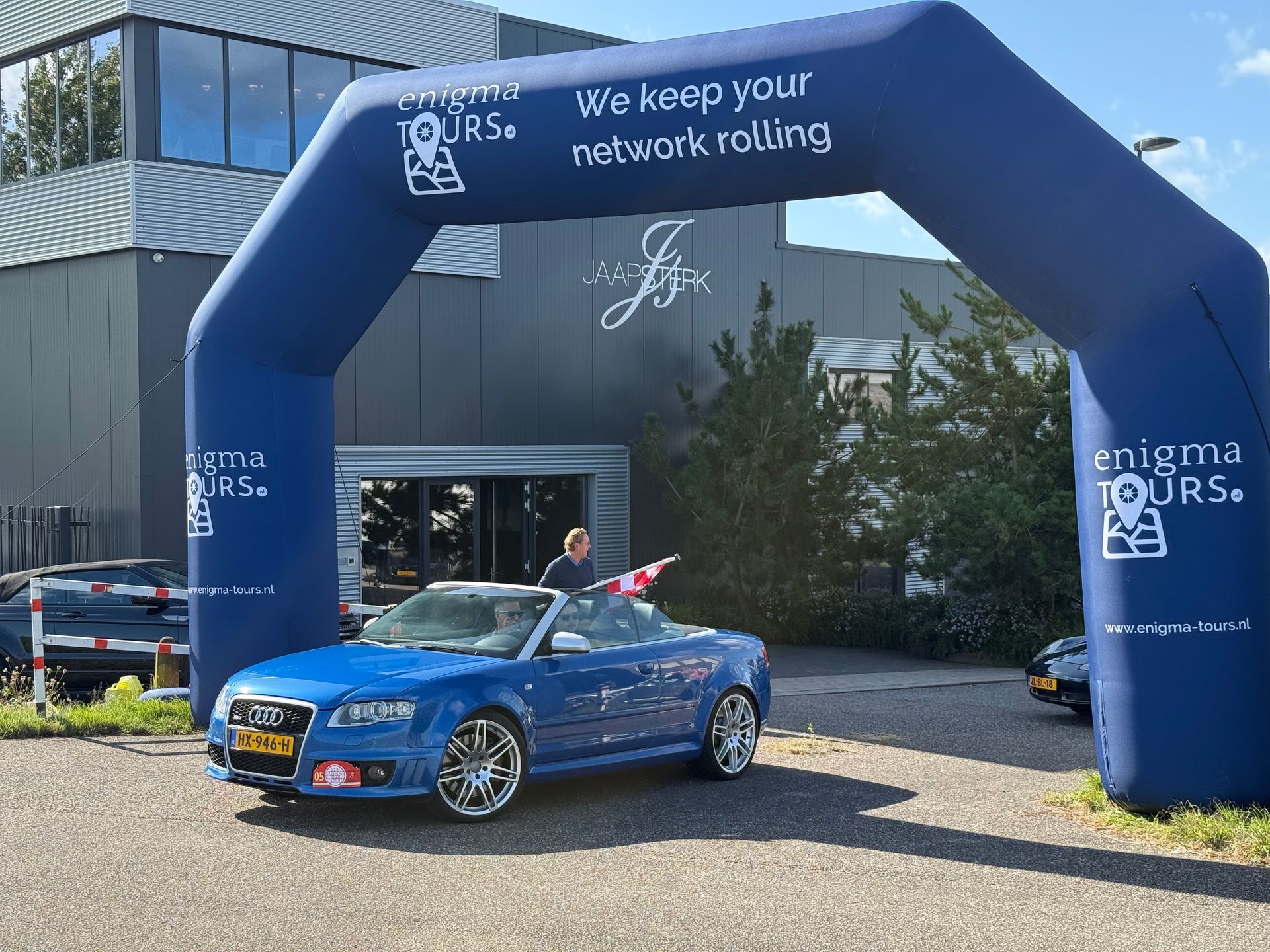 Een blauwe cabriolet rijdt door een opblaasbare boog voor Enigma Tours.