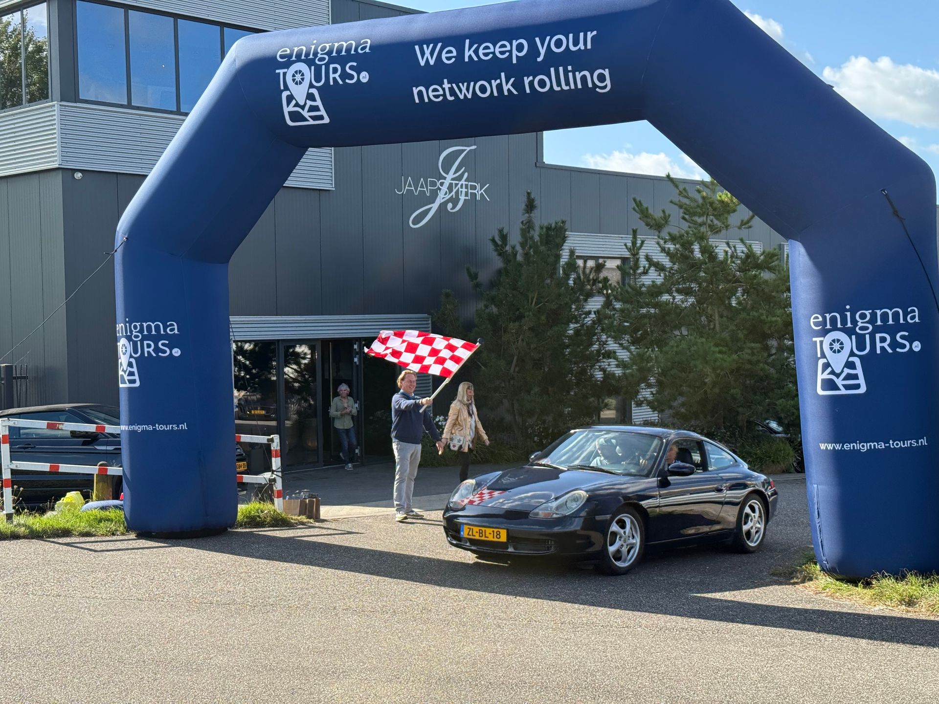 Een donkerblauwe Porsche onder een opblaasbare boog, toegezwaaid door iemand met een geblokte vlag; een gebouw is op de achtergrond te zien.