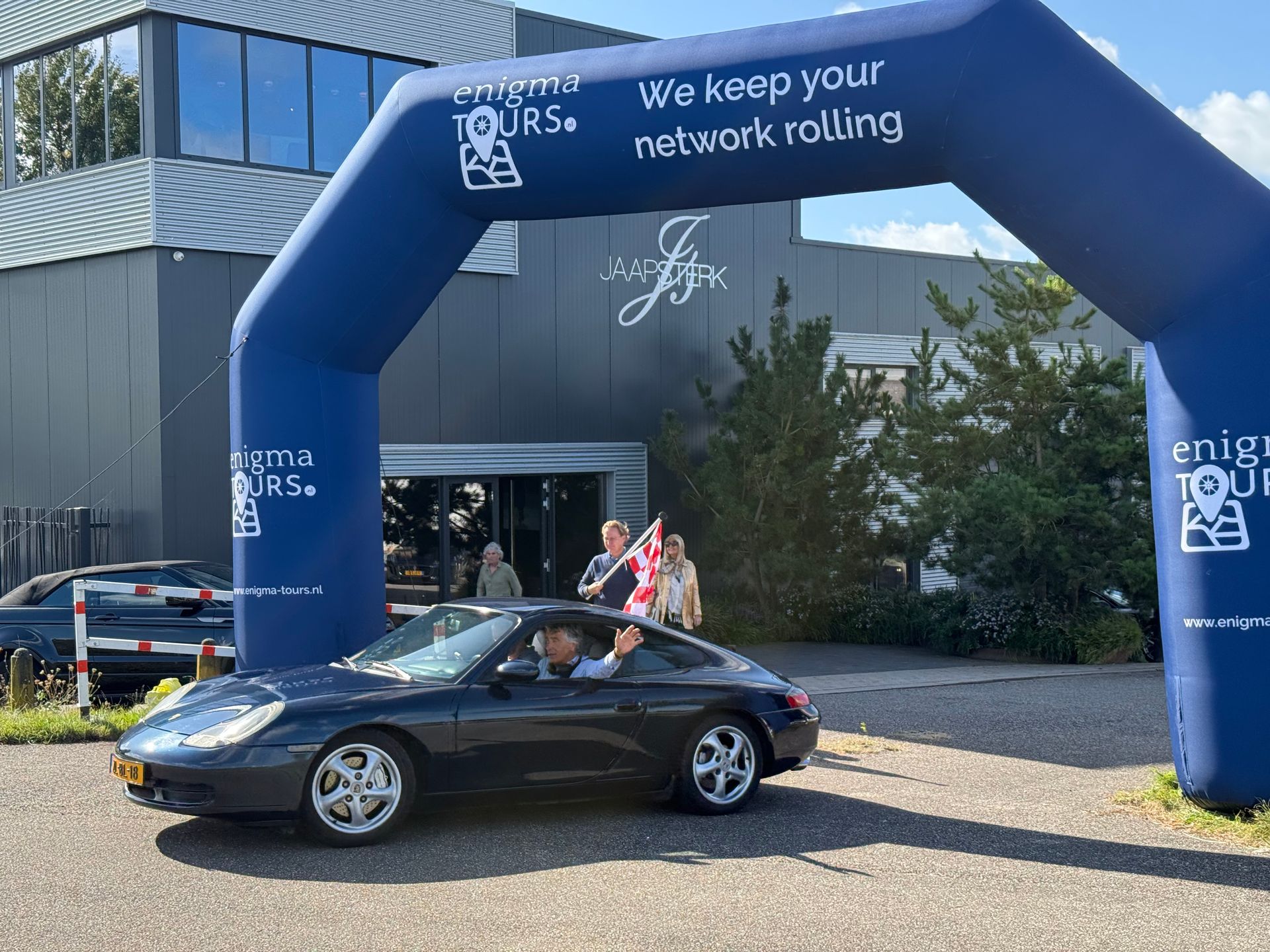 Een donkere Porsche rijdt onder een blauwe boog door, met mensen voor een gebouw.