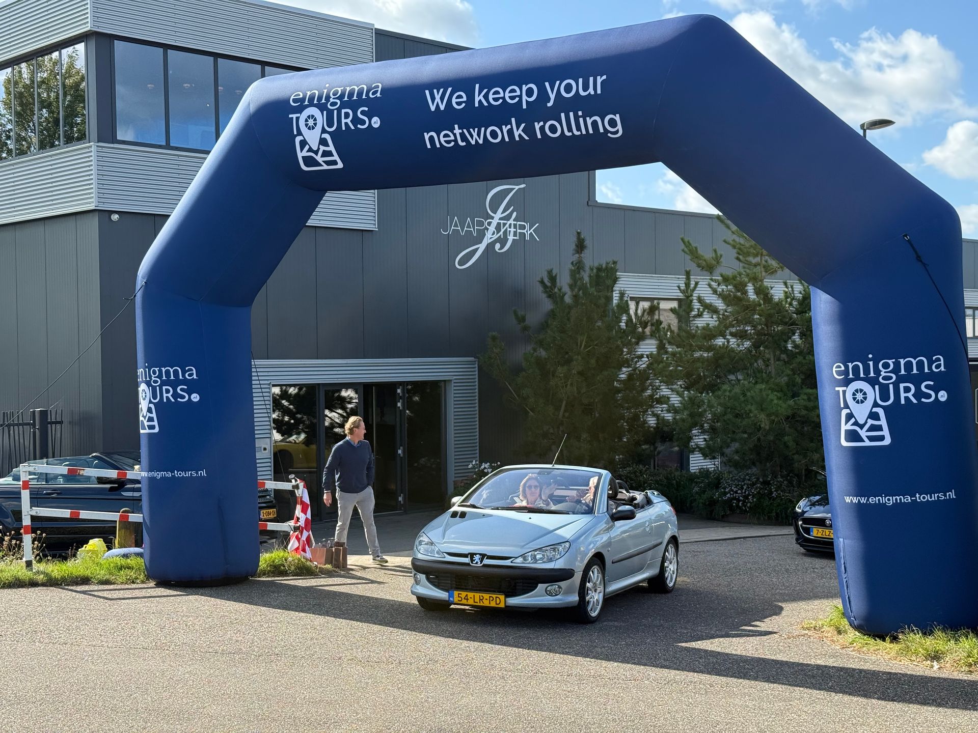 Een cabriolet rijdt door een opblaasbare boog. Op het bord staat