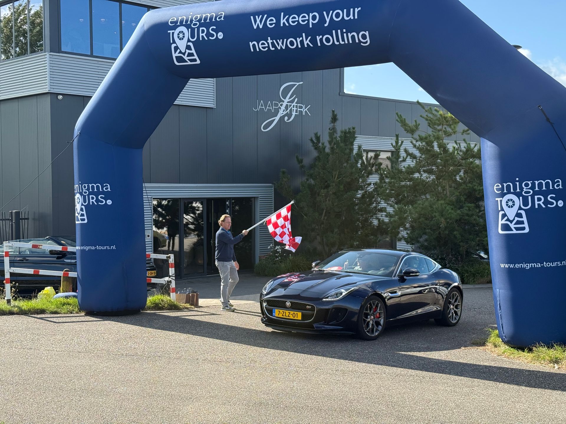 Een man zwaait met een geblokte vlag terwijl een donkerblauwe auto onder een opblaasbare boog doorrijdt, met een gebouw op de achtergrond.