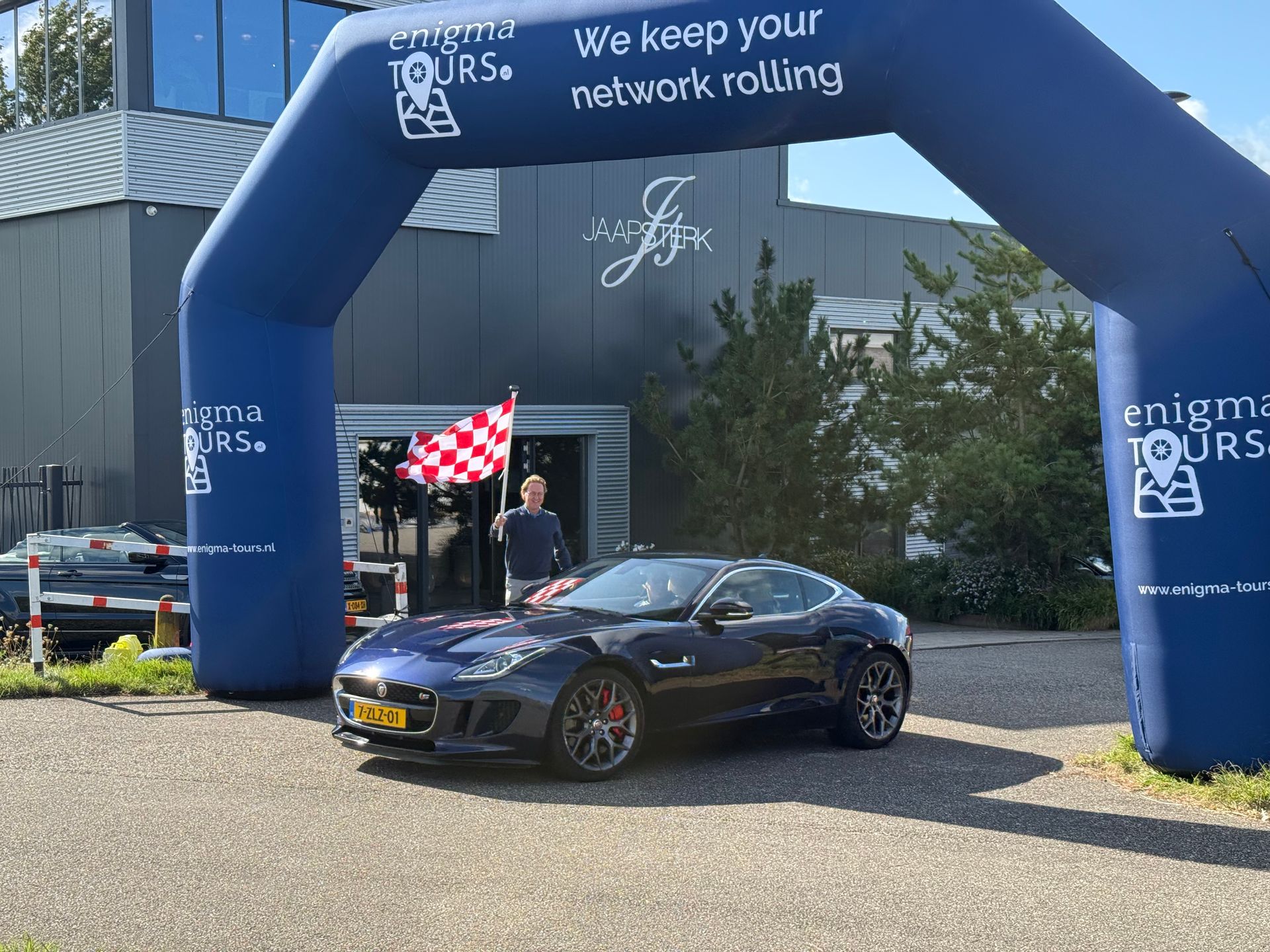 Een blauwe Jaguar sportwagen onder een boog, een man met een geblokte vlag, voor een gebouw met een blauwe boog.