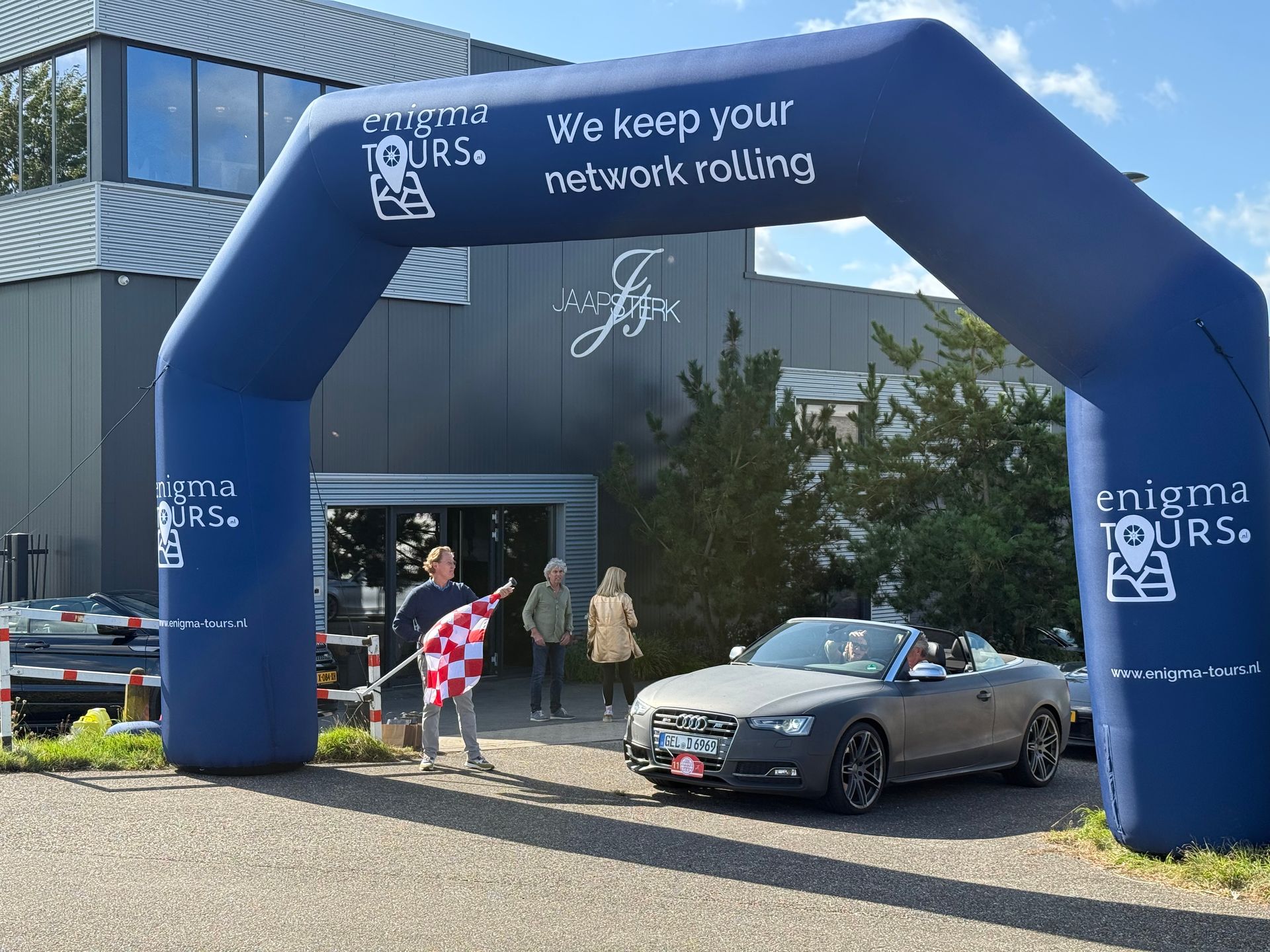 Een persoon zwaait met een geblokte vlag naast een grijze cabriolet onder een boog van