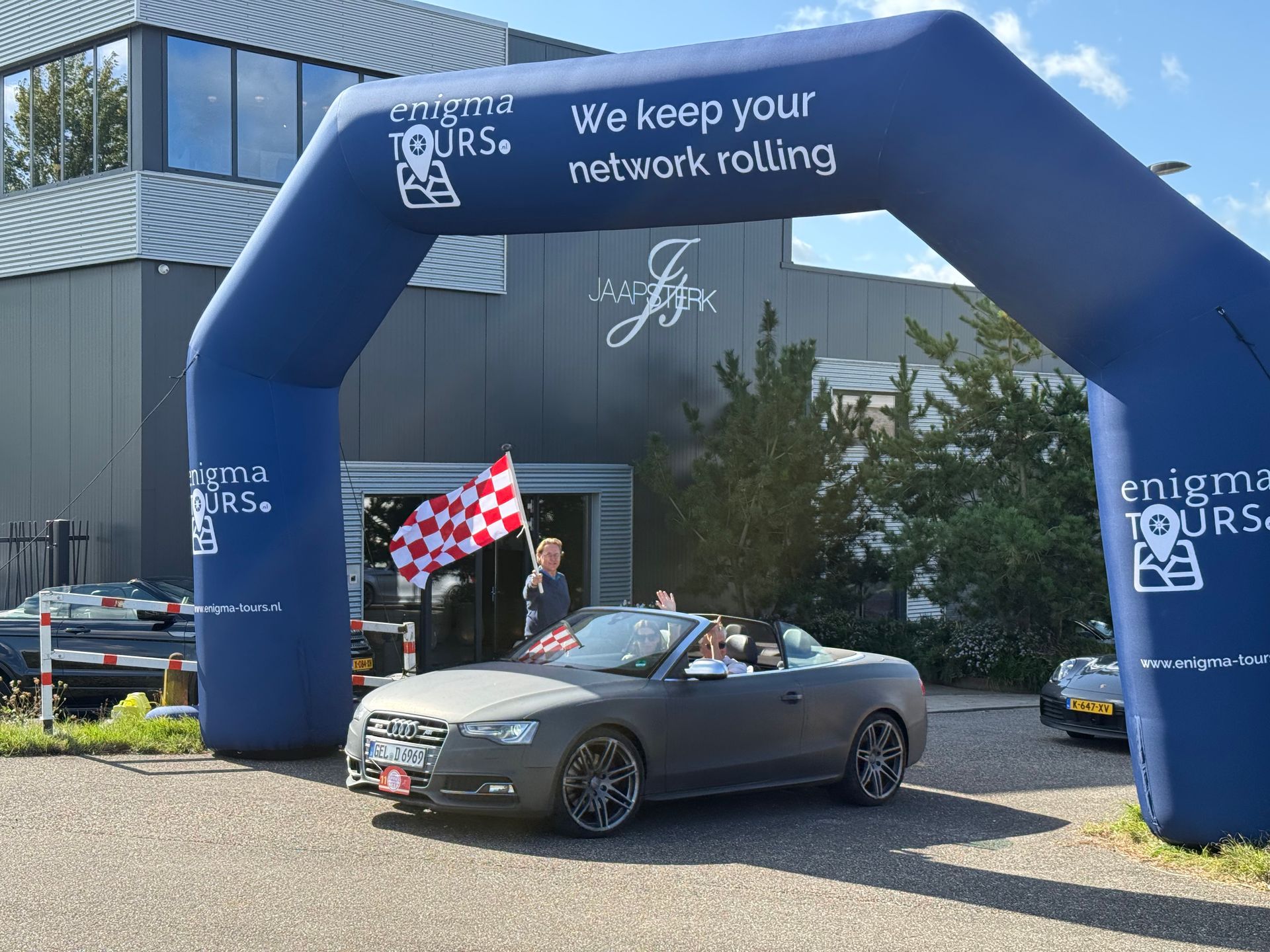 Een grijze cabriolet rijdt onder een blauwe boog door met de tekst