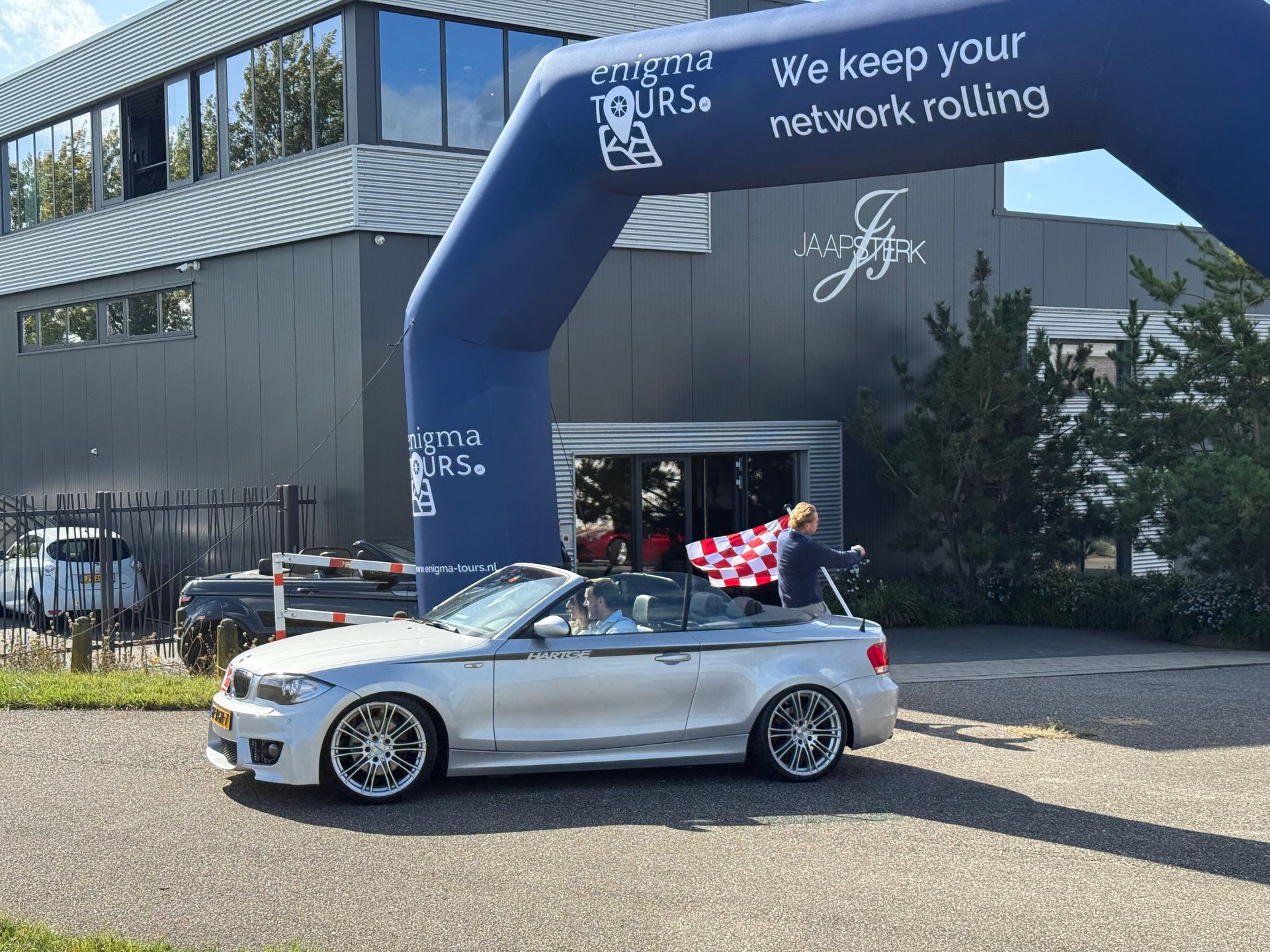 Een zilverkleurige cabriolet verlaat een gebouw onder een boog. Iemand zwaait met een vlag.
