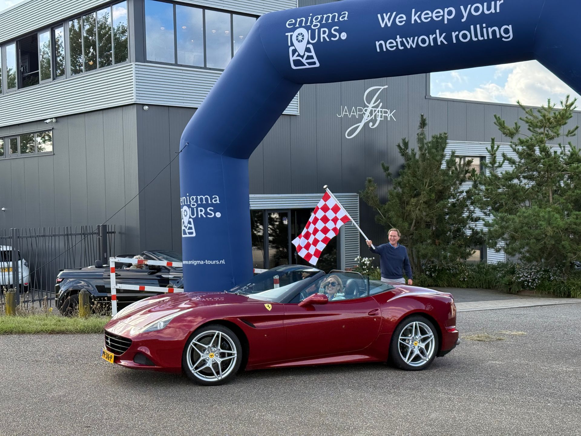 Een man zwaait met een geblokte vlag naast een rode Ferrari cabriolet onder een boog met het opschrift