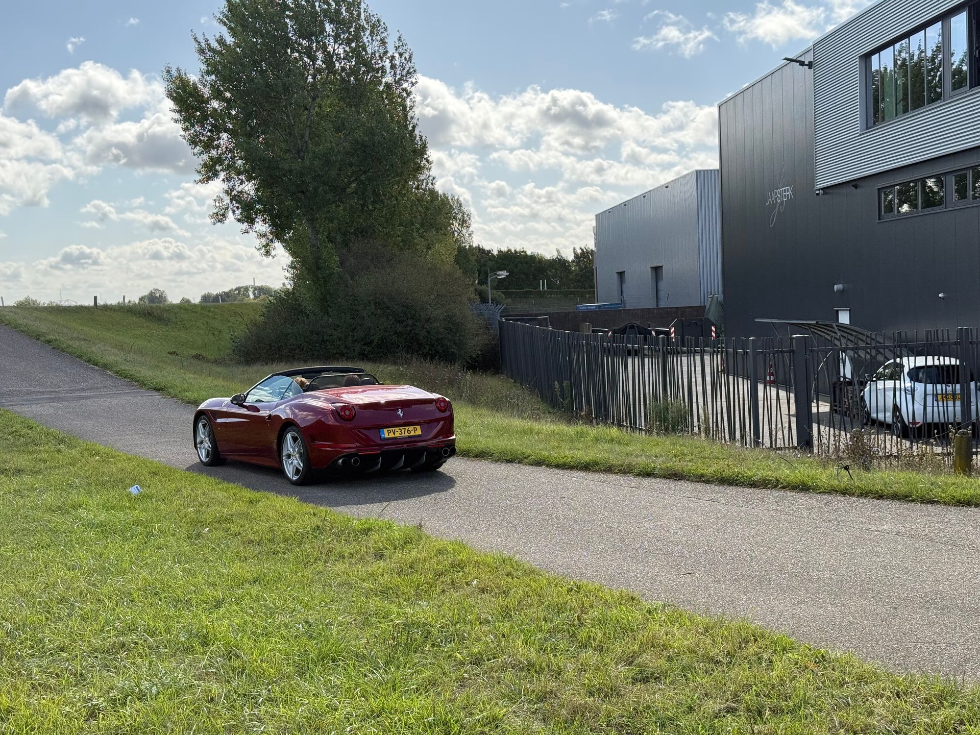 Een rode cabriolet rijdt op een zonnige dag over een weg langs een grasveld en een modern gebouw.