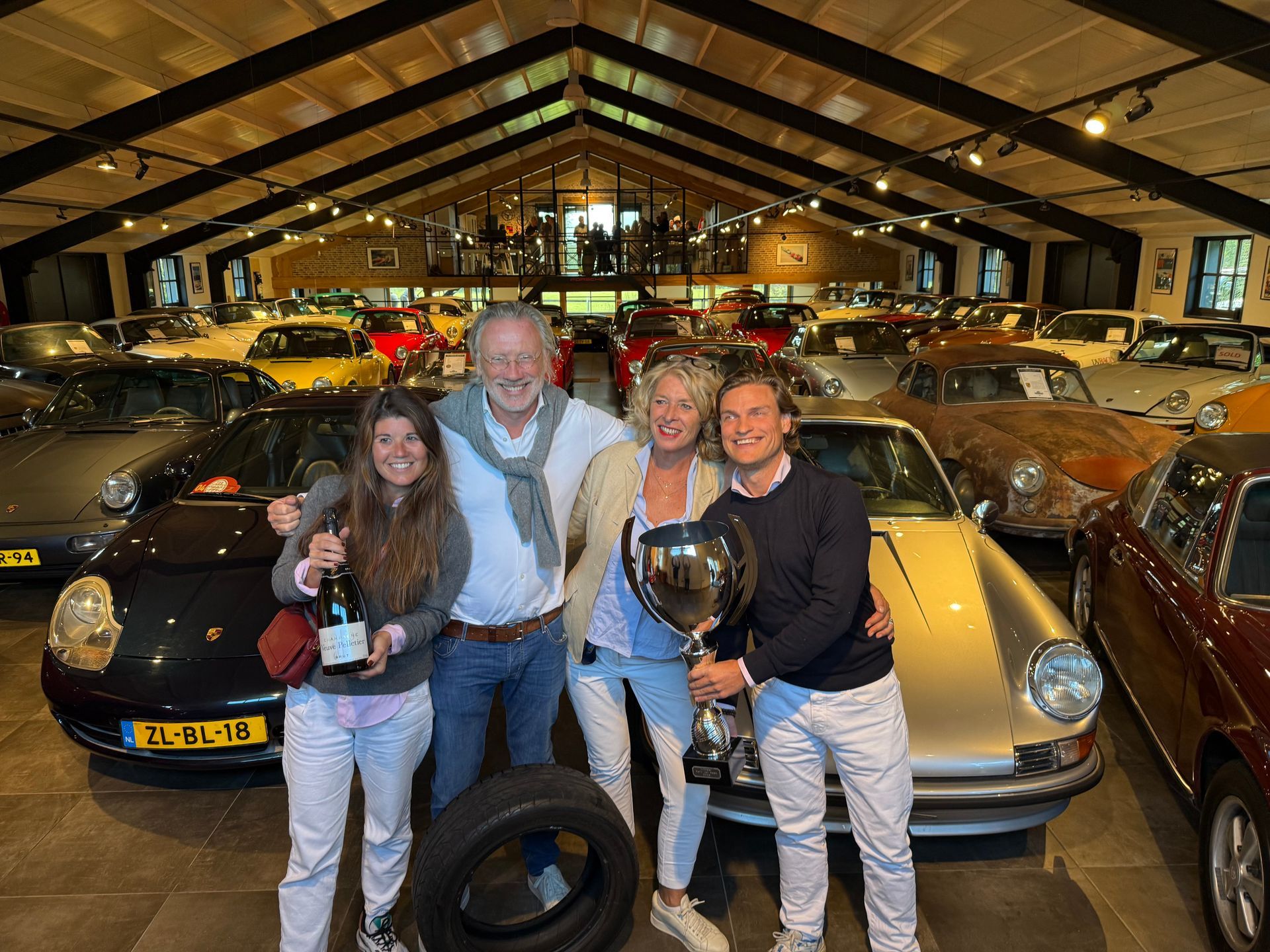 Vier mensen poseren met trofeeën in een showroom voor klassieke auto's.
