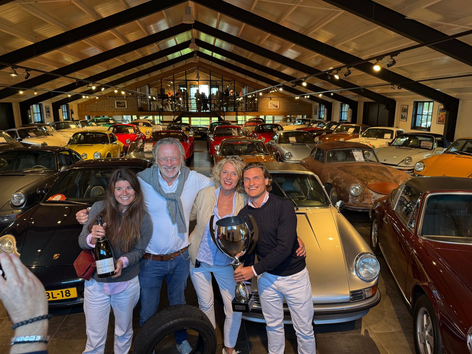Vier mensen vieren feest met een trofee en champagne in een autoshowroom vol oldtimers.