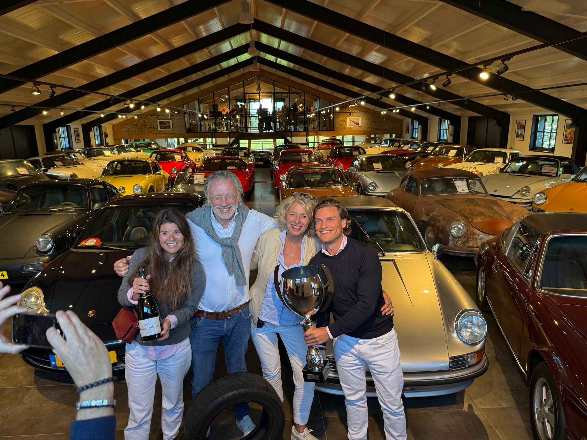 Een groep met een trofee, omringd door klassieke auto's in een grote garage. De mensen glimlachen.