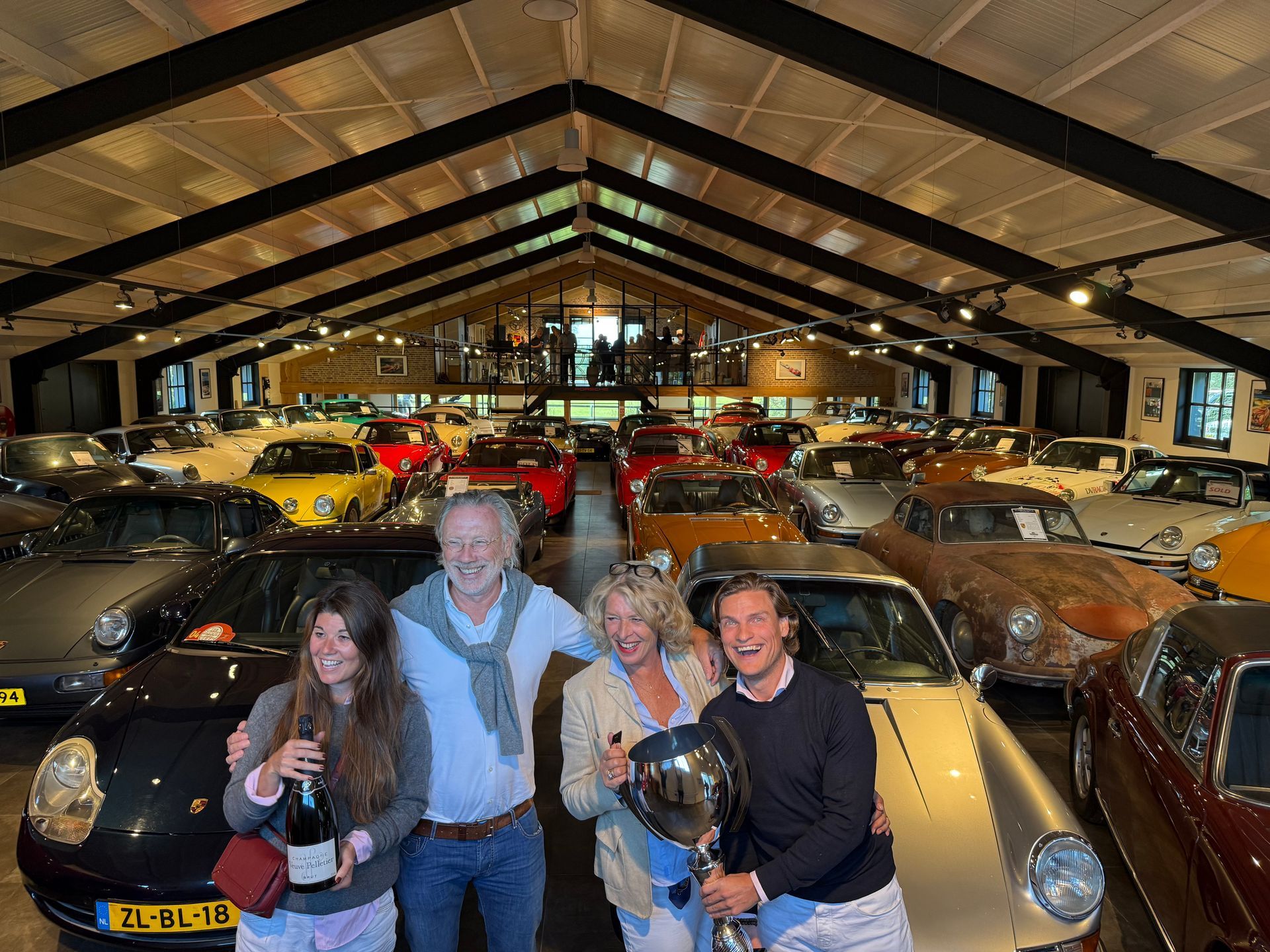 Mensen poseren met auto's in een grote garage; de een houdt een trofee vast, de ander champagne.