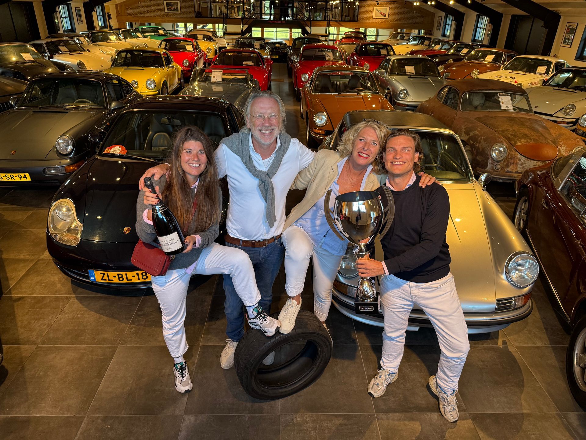 Vier mensen vieren feest met een trofee en champagne te midden van een showroom vol klassieke auto's.