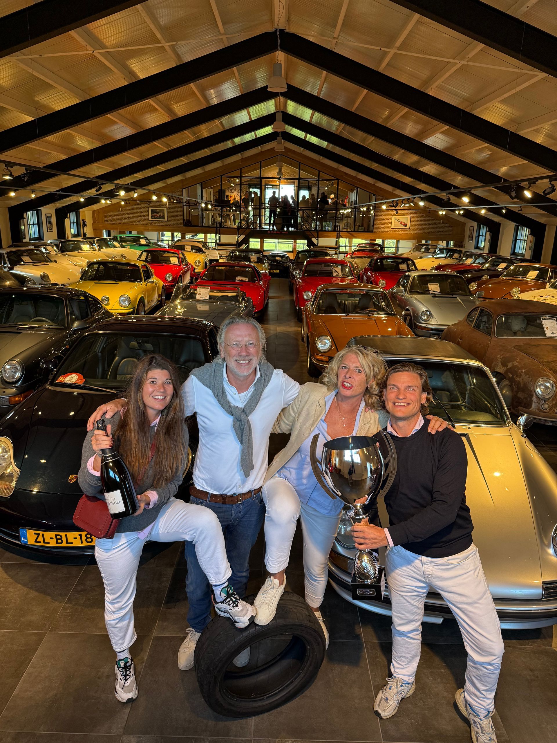 Vier mensen poseren met een trofee, champagne en een band in een autoshowroom vol klassieke auto's.