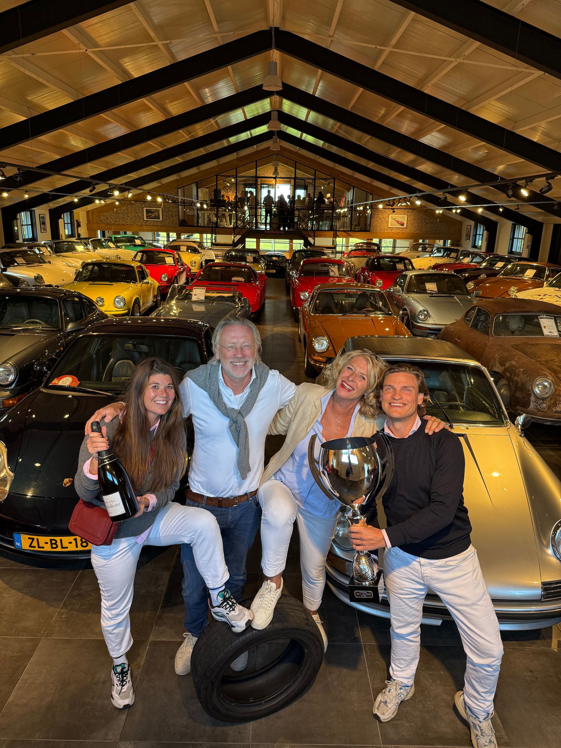 Vier mensen vieren feest met champagne en een trofee in een garage vol klassieke auto's.