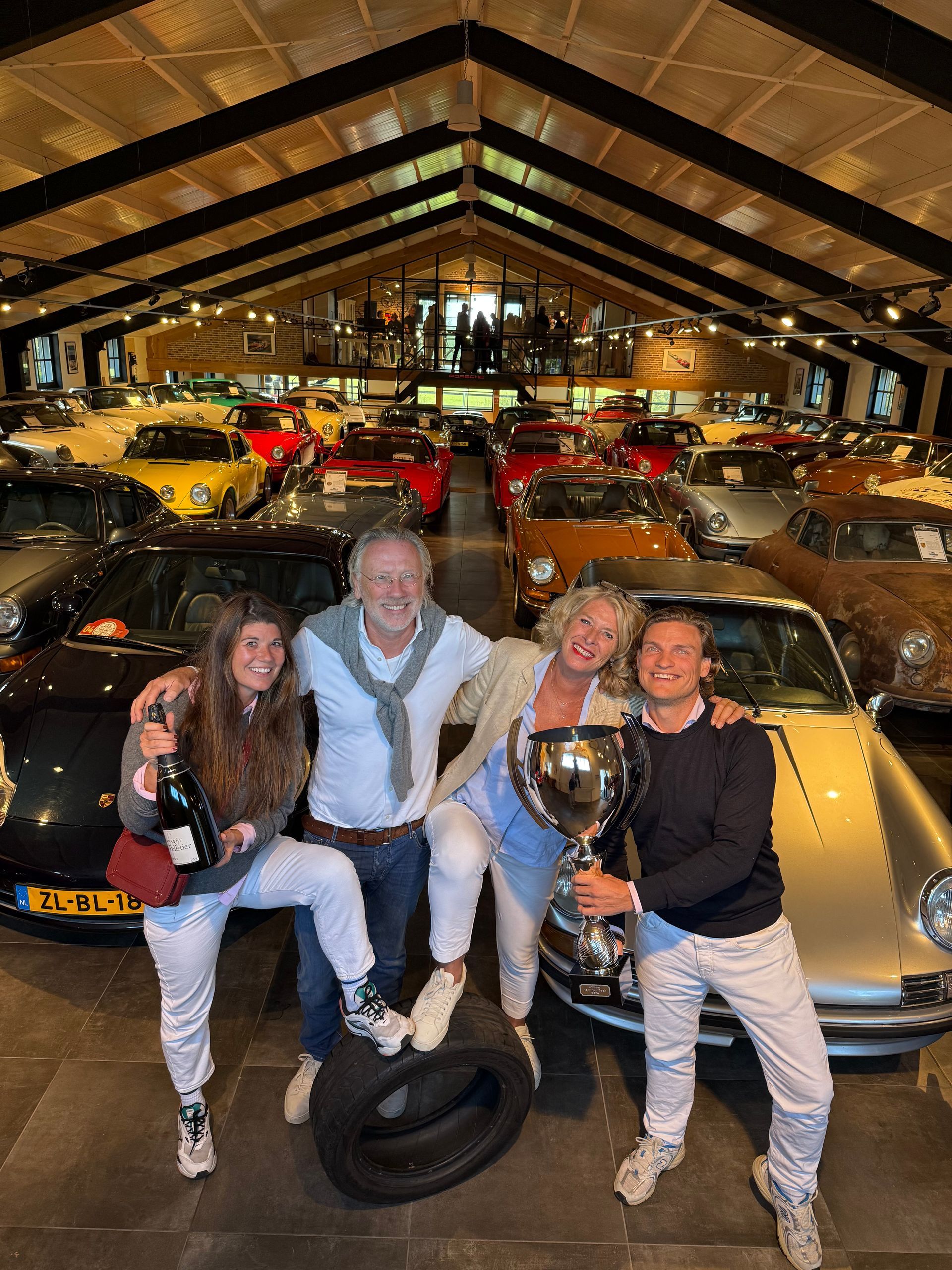 Vier mensen vieren feest met een champagnefles en een trofee in hun handen in een autoshowroom vol Porsches.