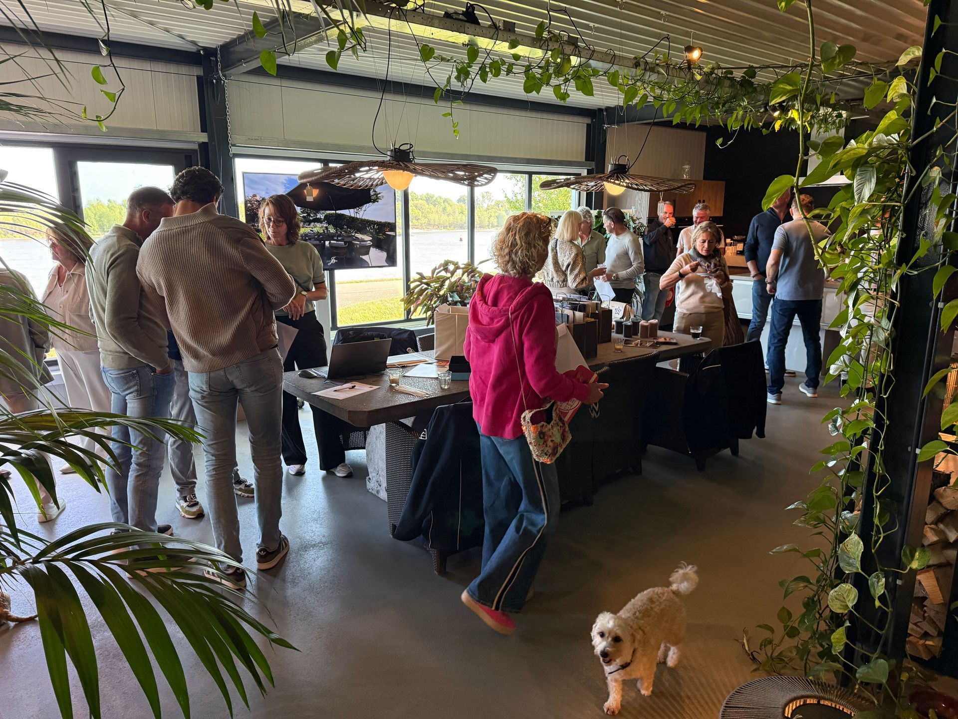 Mensen op een evenement, verzameld rond een tafel met diverse voorwerpen, planten op de voorgrond en ramen op de achtergrond.