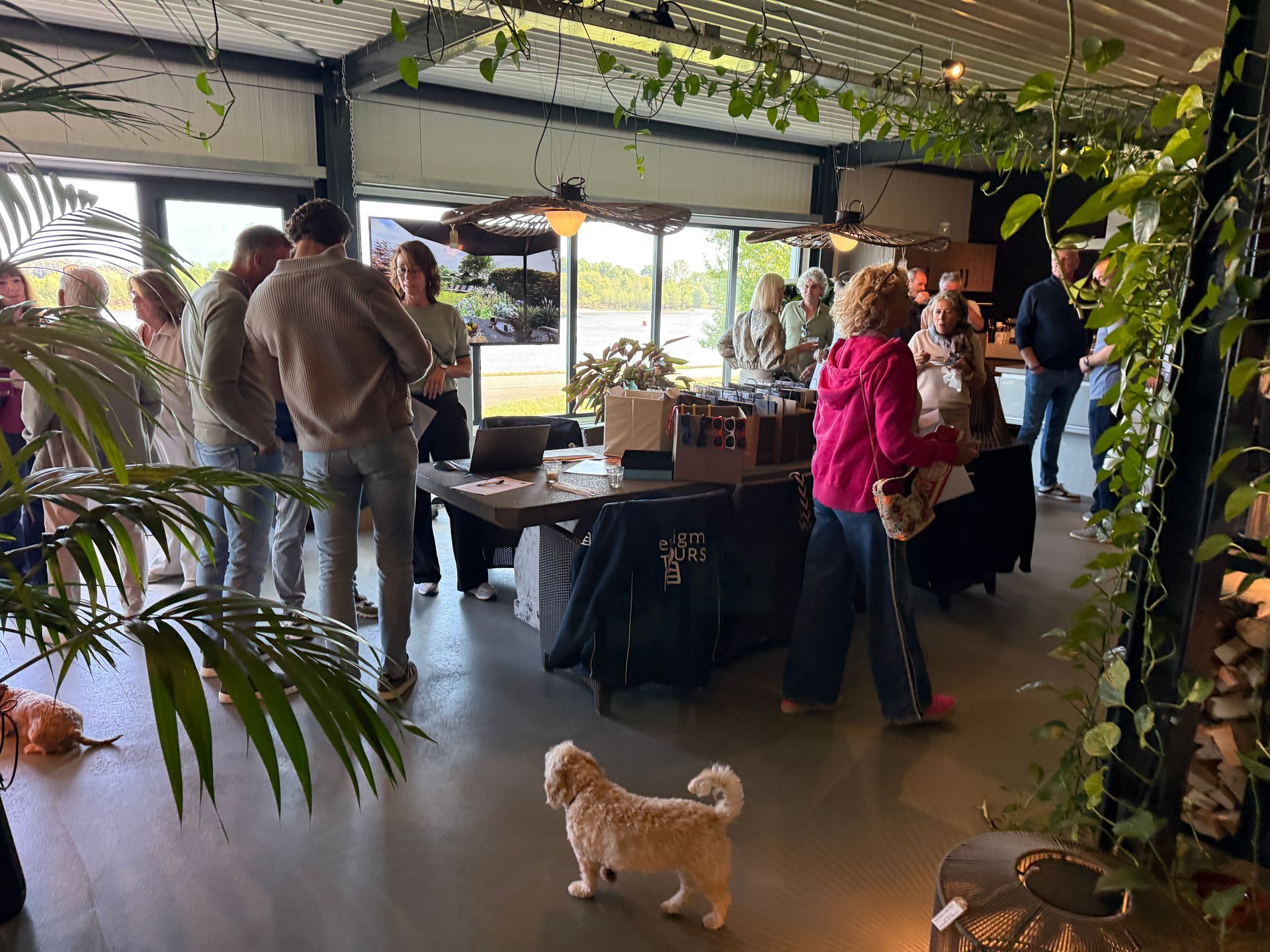 Mensen komen samen in een modern gebouw met planten; sommigen kijken naar een tafel met koopwaar. Een hond staat op de voorgrond.