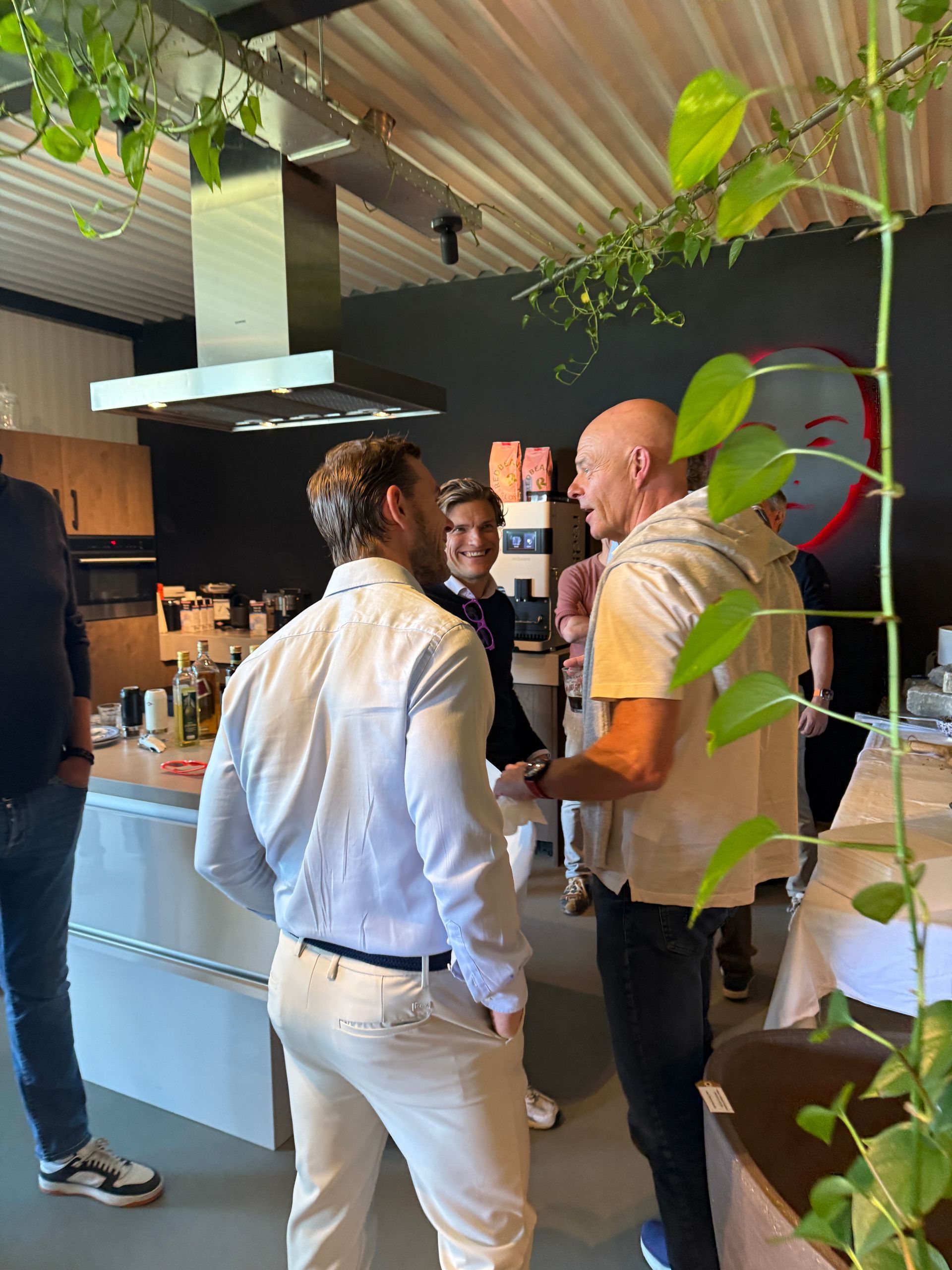 Twee mannen in gesprek in een moderne keuken. De ene draagt ​​een licht overhemd, de andere een licht shirt en een donkere broek.