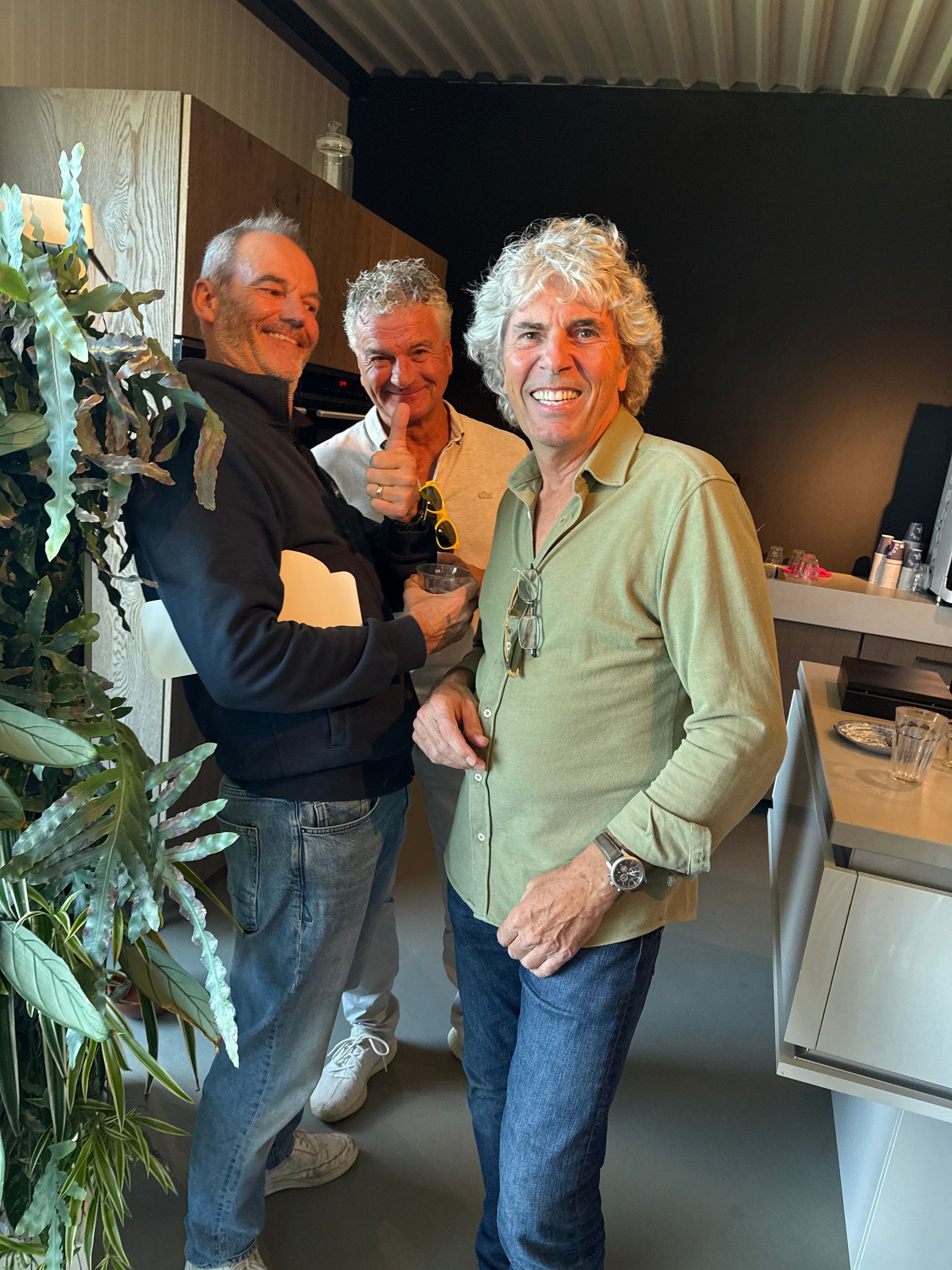 Drie mannen glimlachen binnen; een houdt een drankje vast, een ander steekt zijn duim omhoog.