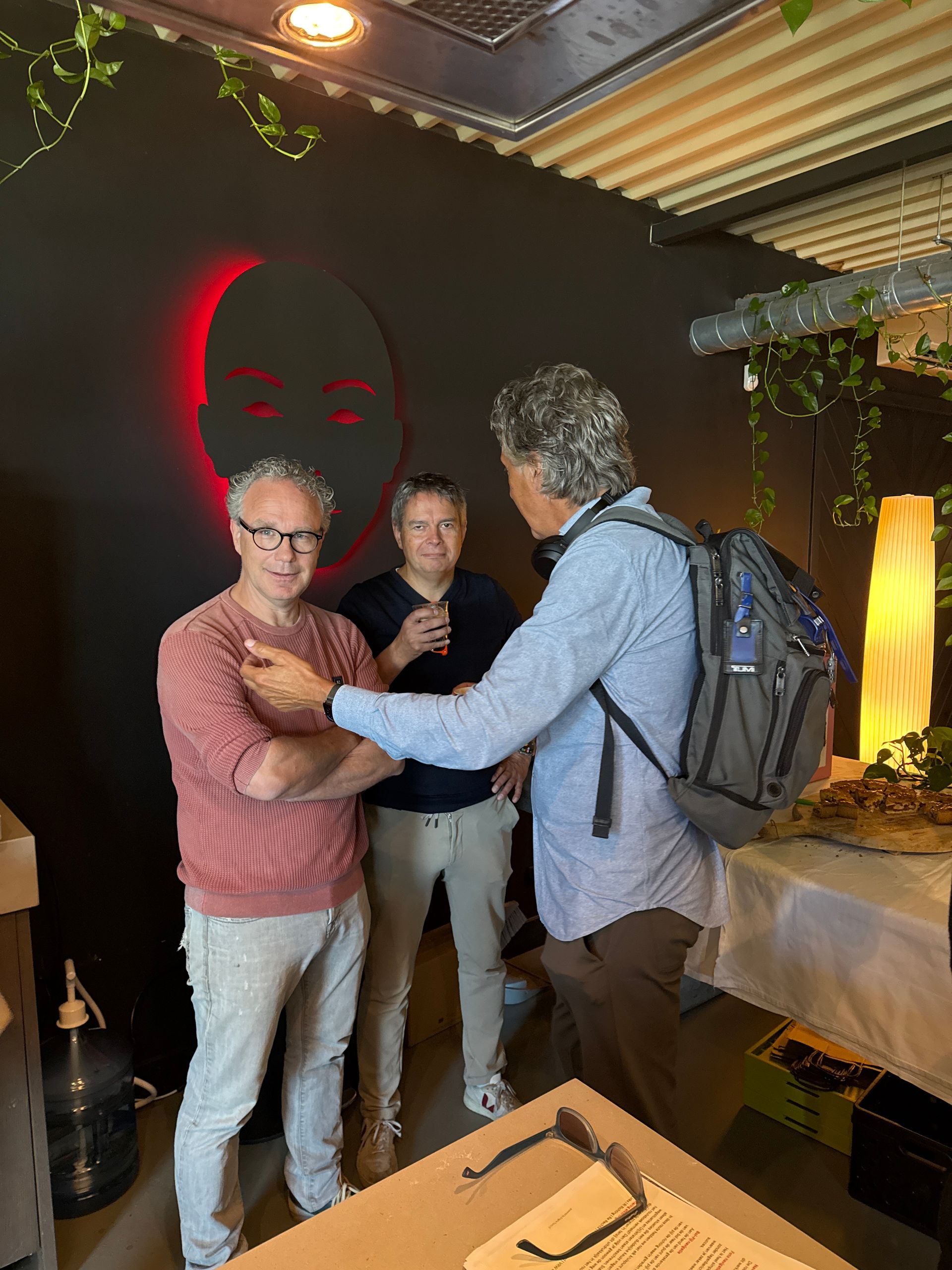 Drie mannen staan ​​binnen, naast een zwarte muur met een rood gezichtssilhouet. Een van hen gebaart.