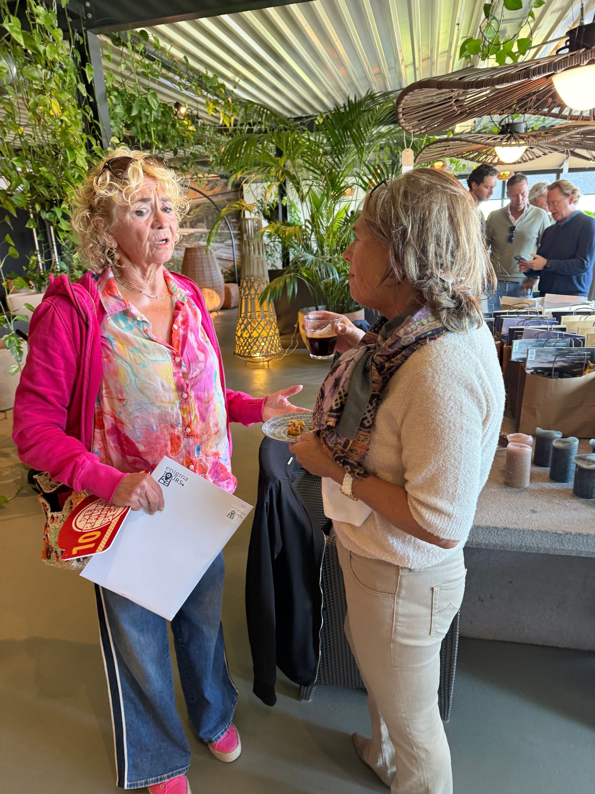 Twee vrouwen in gesprek tijdens een evenement, een van hen met een drankje in de hand. Omringd door groen, tafels en mensen.