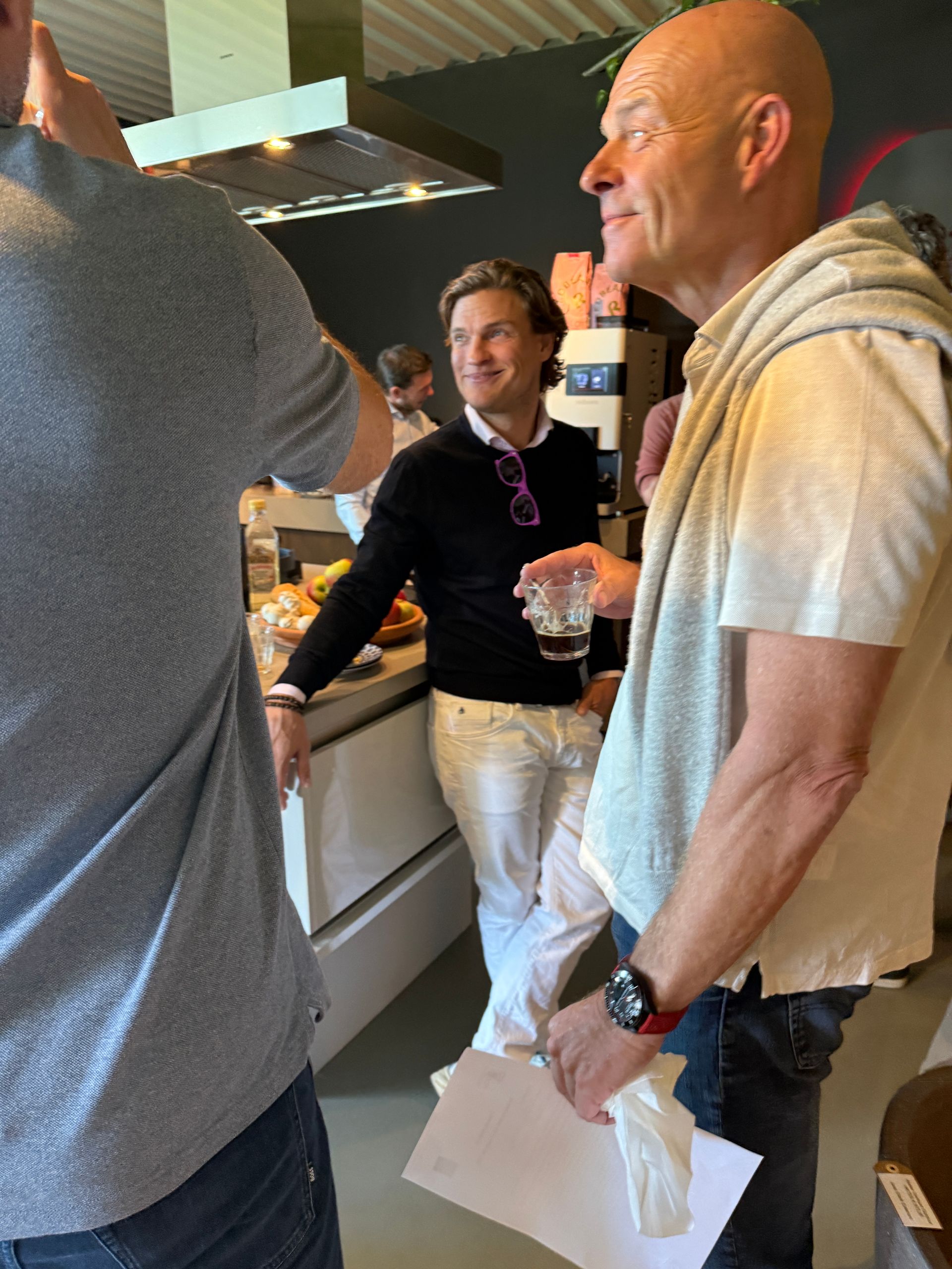 Drie mannen in een keuken; een houdt een drankje vast, een ander maakt een foto. De man in het midden glimlacht.