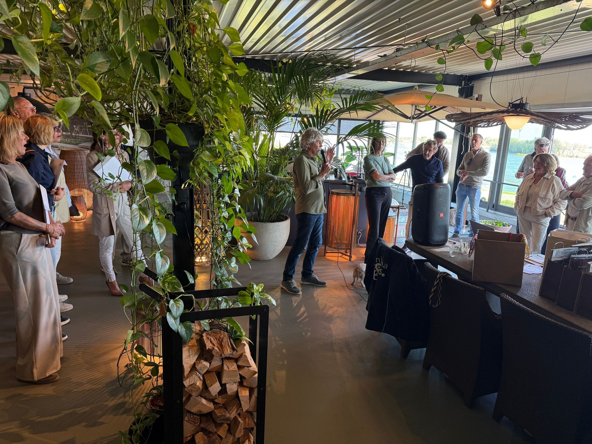 Mensen op een evenement in een zaal vol planten. Een man spreekt, anderen luisteren.