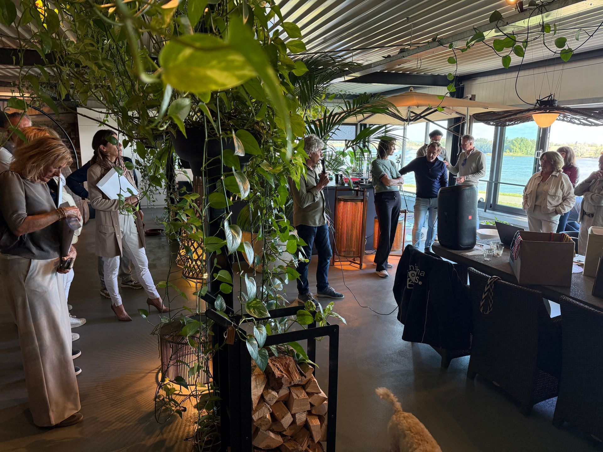 Mensen op een evenement met planten en uitzicht op water, één persoon spreekt vlakbij een luidspreker, binnen.