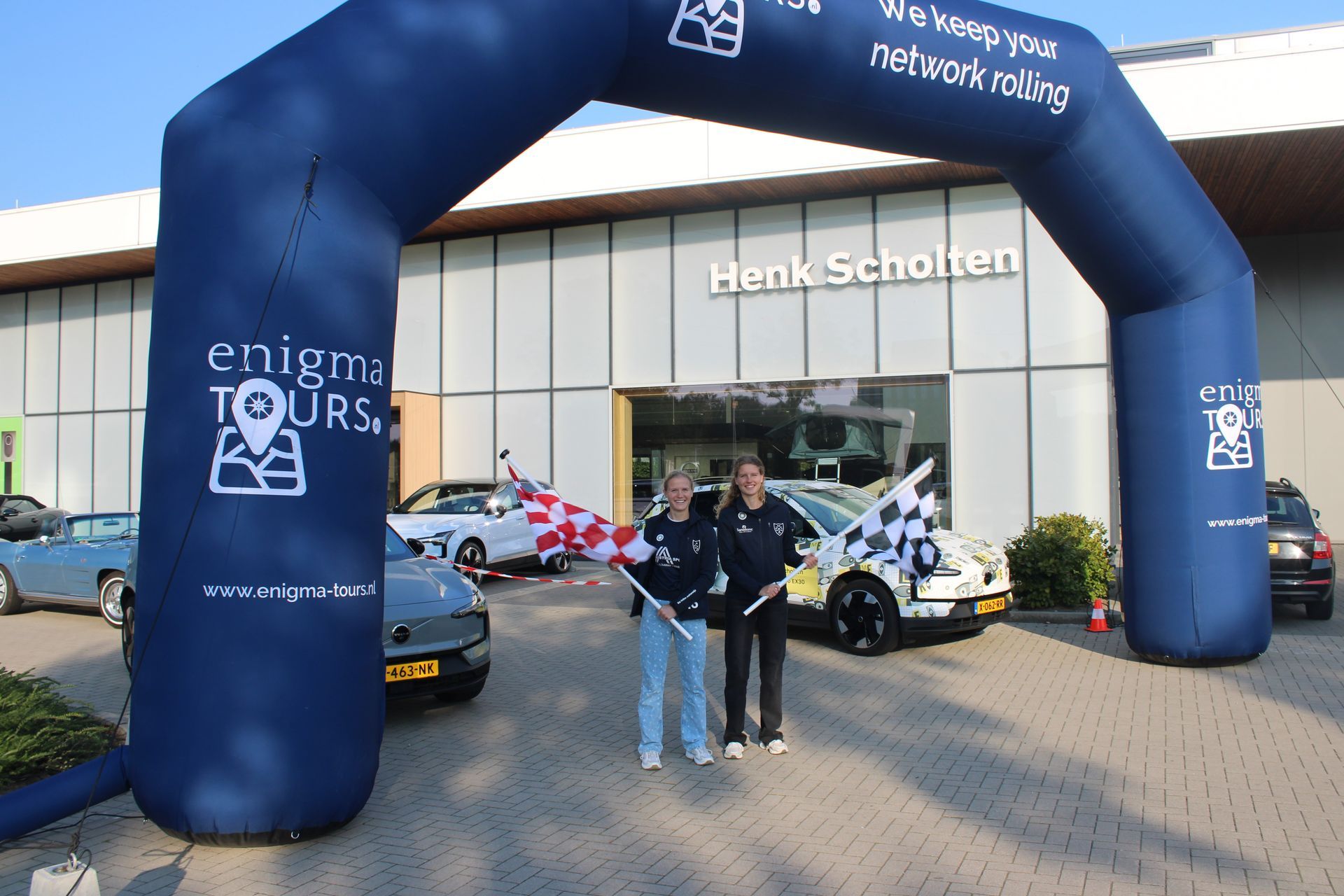Twee mensen staan ​​onder een opblaasbare boog bij autodealer Henk Scholten. Er staan ​​auto's in de buurt geparkeerd.