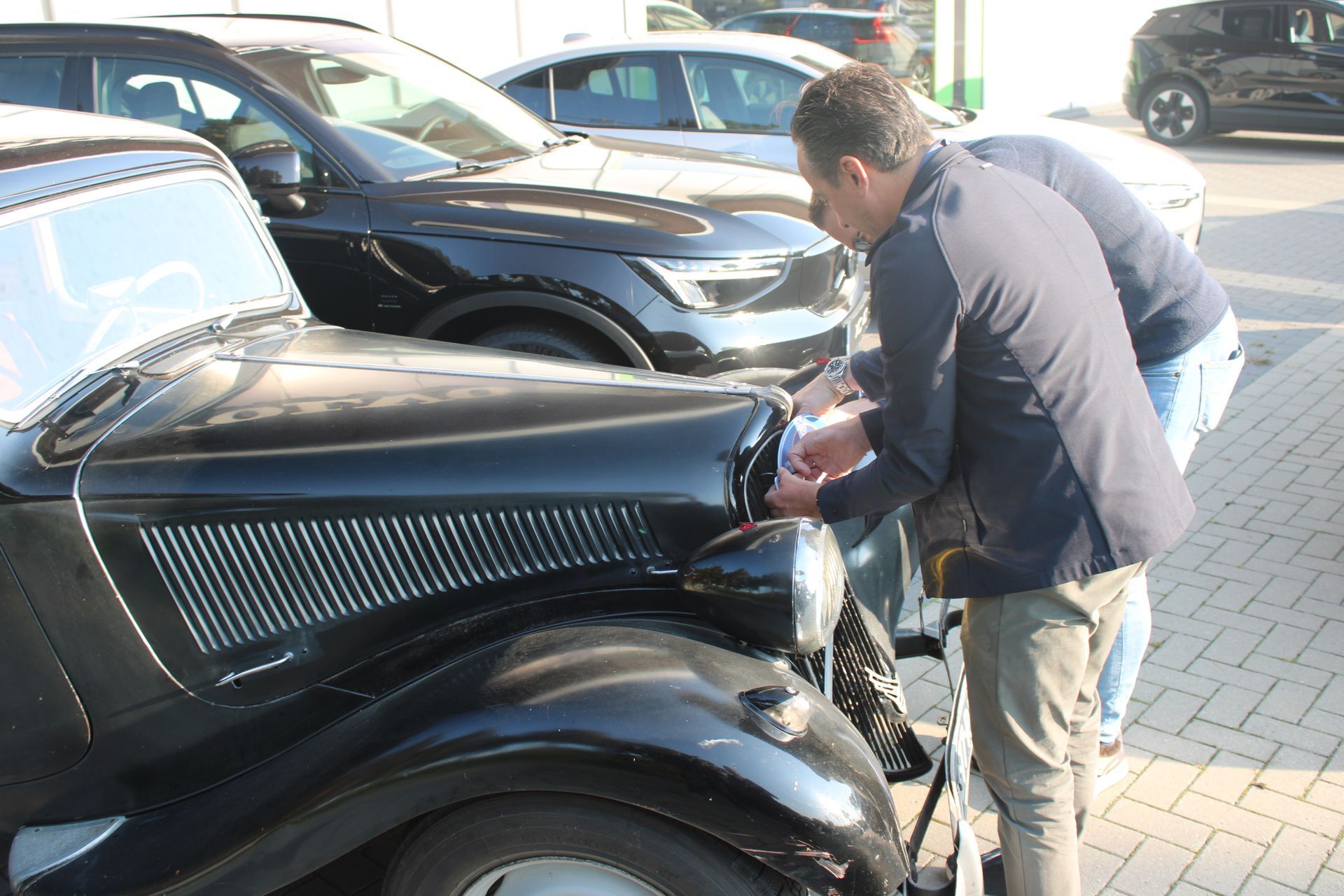 Twee mannen inspecteren de koplamp van een zwarte oldtimer. Op de achtergrond staan ​​andere auto's geparkeerd.