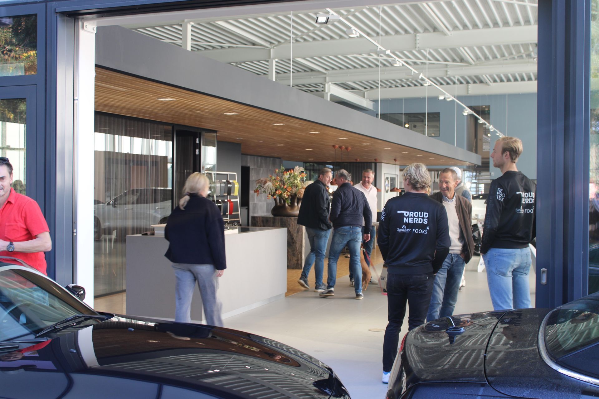 Mensen bij een autodealer, sommigen kijken naar binnen. Zwarte auto's op de voorgrond, open ingang, modern interieur.