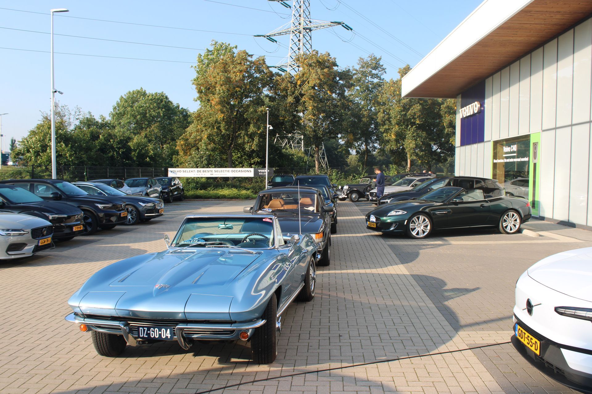 Een blauwe klassieke cabriolet staat op een parkeerplaats met andere voertuigen, vlakbij een gebouw, op een zonnige dag.
