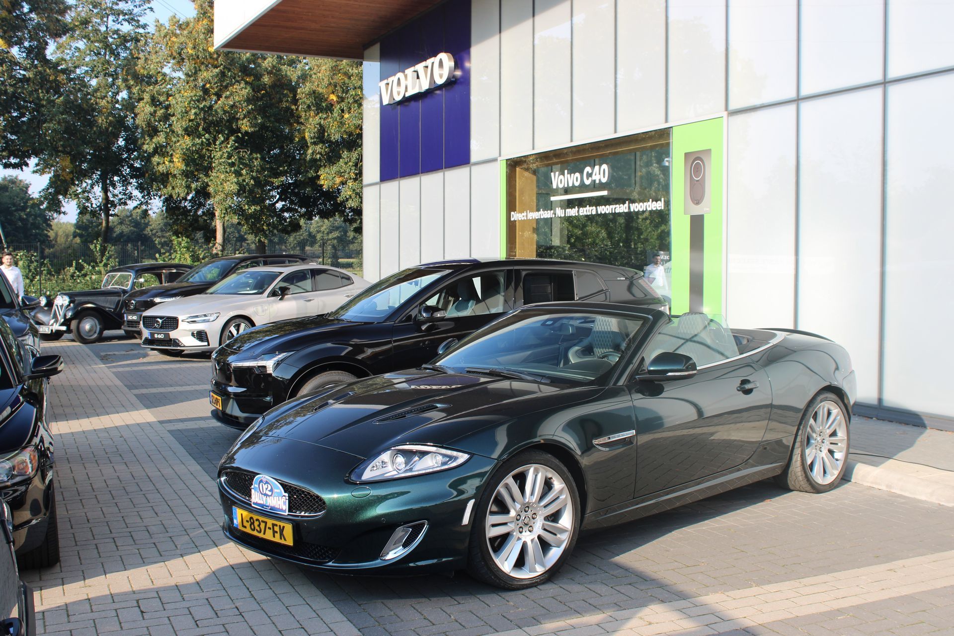 Een groene Jaguar cabriolet staat geparkeerd voor een Volvo-dealer. Op de achtergrond zijn andere auto's, een gebouw en groen te zien.