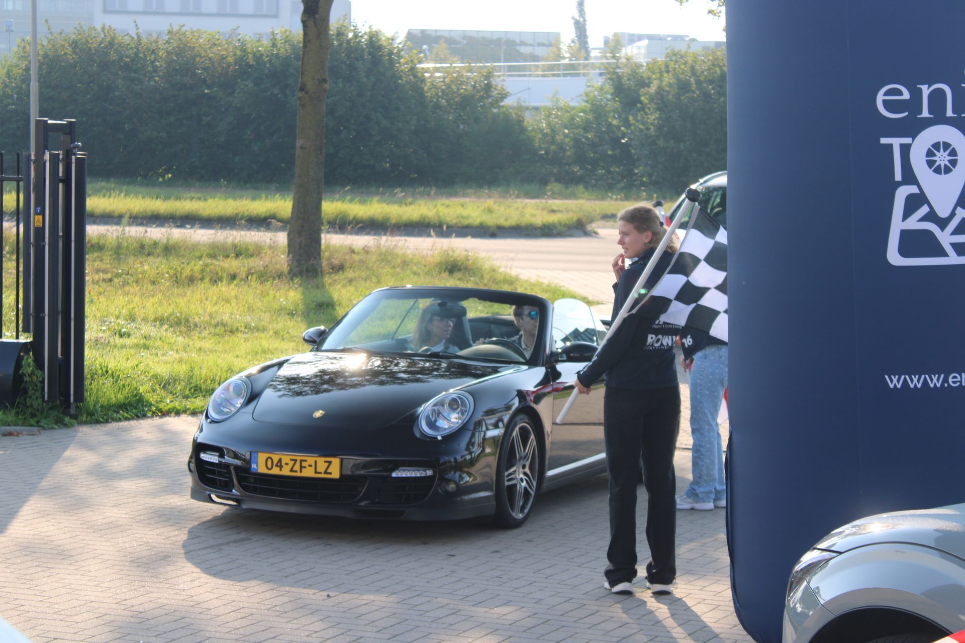 Zwarte Porsche cabriolet met bestuurder; persoon zwaait met geblokte vlag.