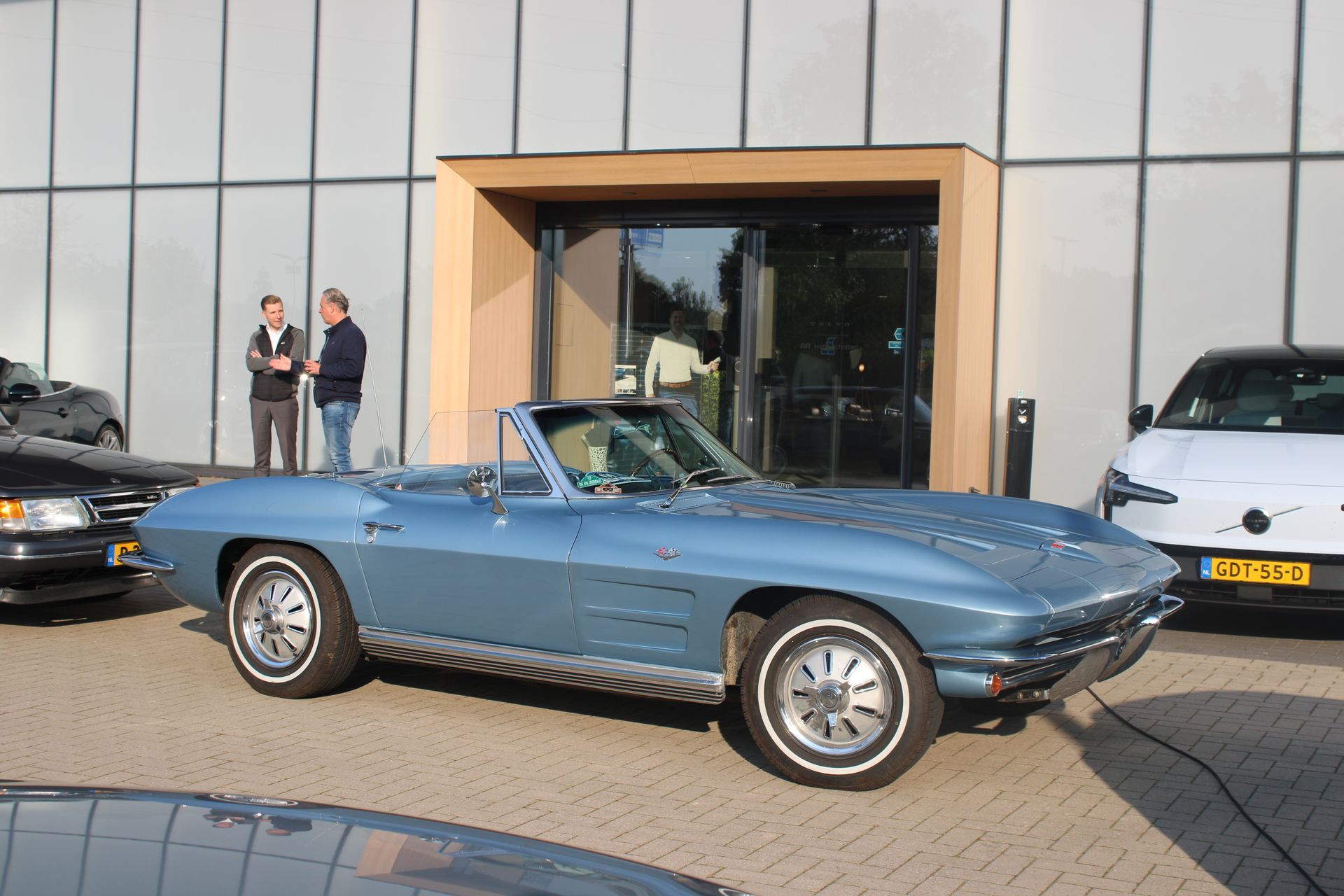 Een blauwe cabriolet staat geparkeerd voor een modern gebouw. ​​Twee mannen staan ​​bij de ingang.